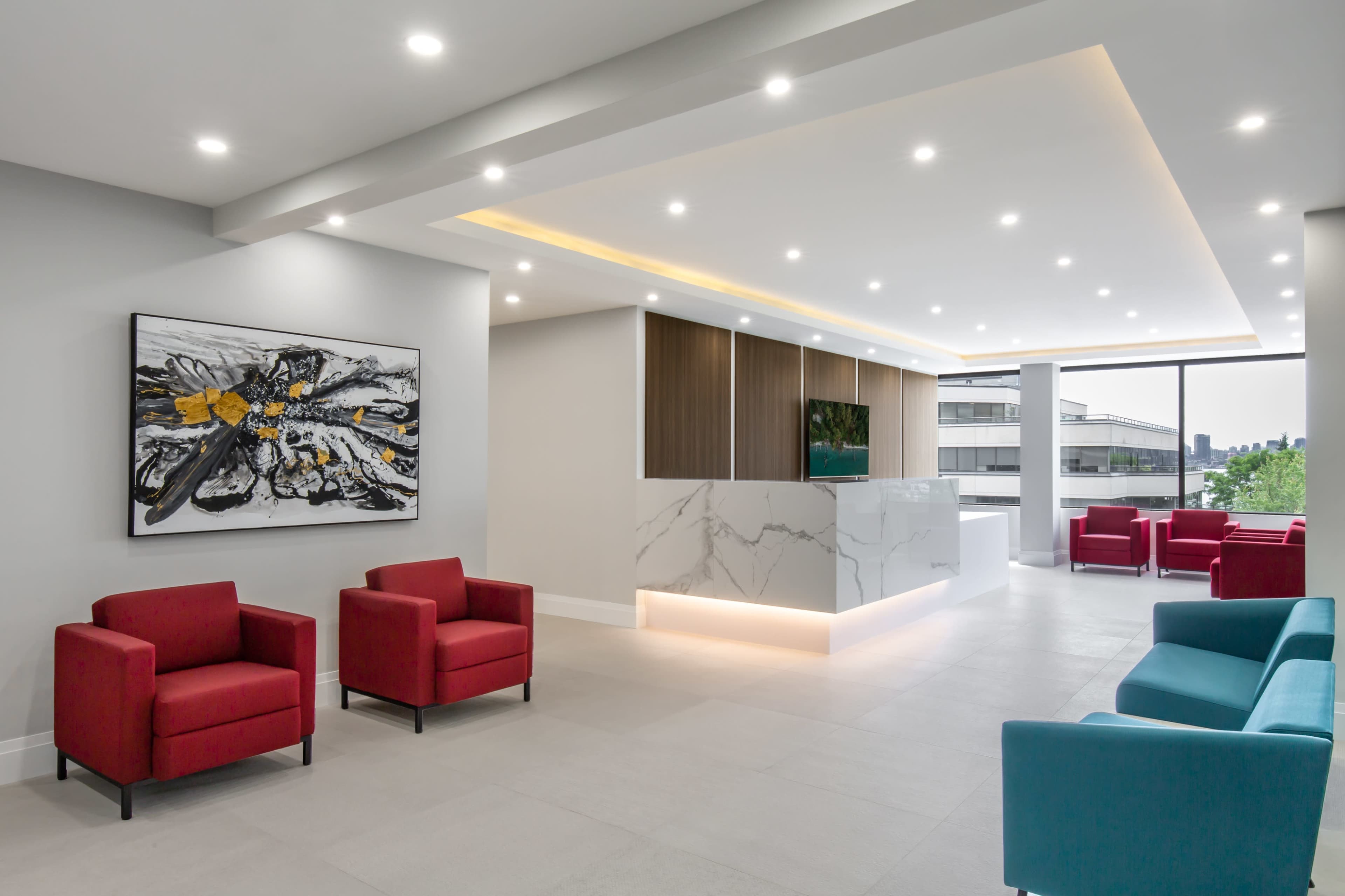A modern reception area features a marble front desk, contemporary seating in red and teal, and large windows overlooking a city landscape.