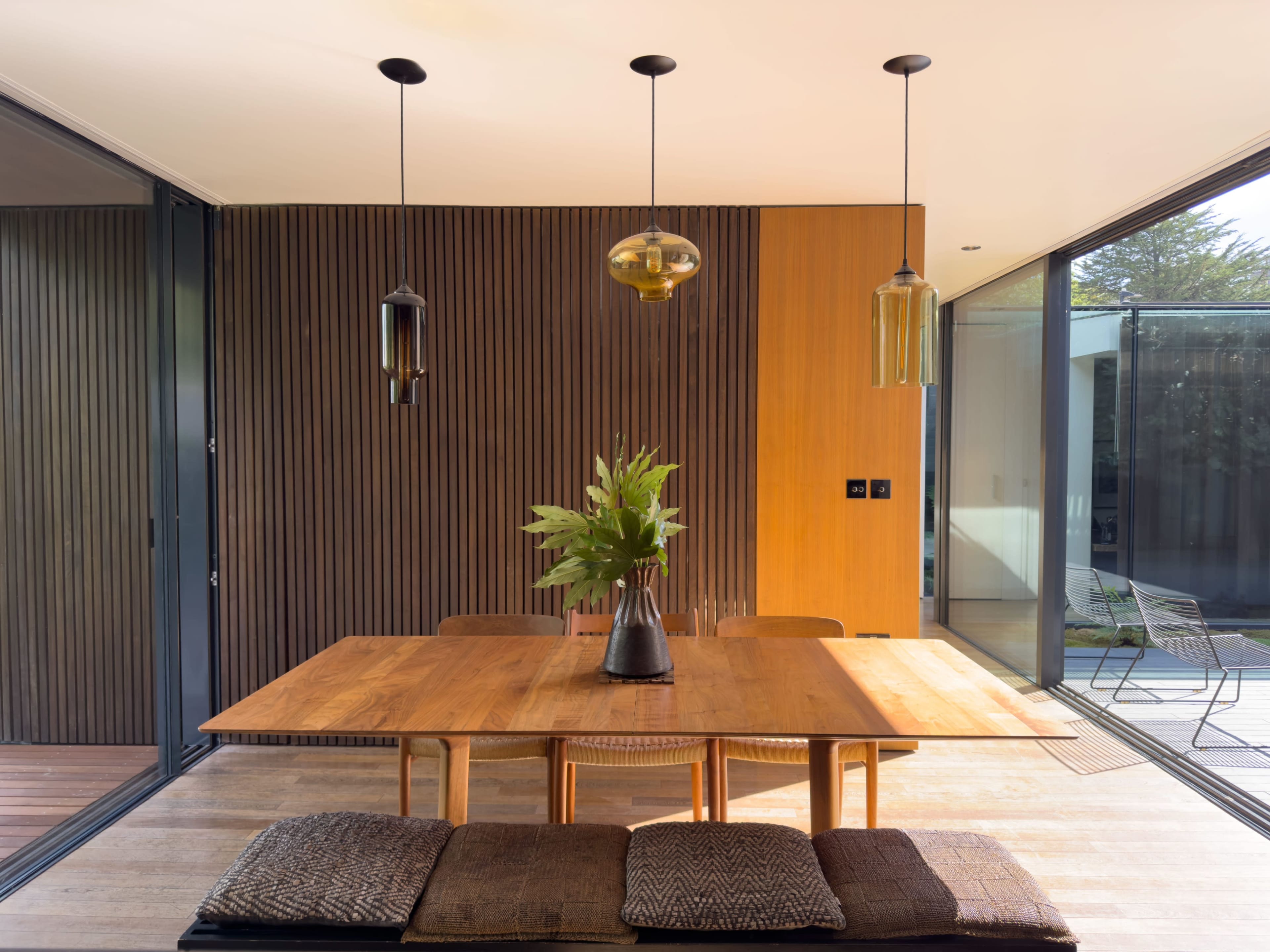 The image features a modern dining area with a wooden table, several pendant lights, and a vase of greenery on the table, framed by large glass windows and wooden paneling.