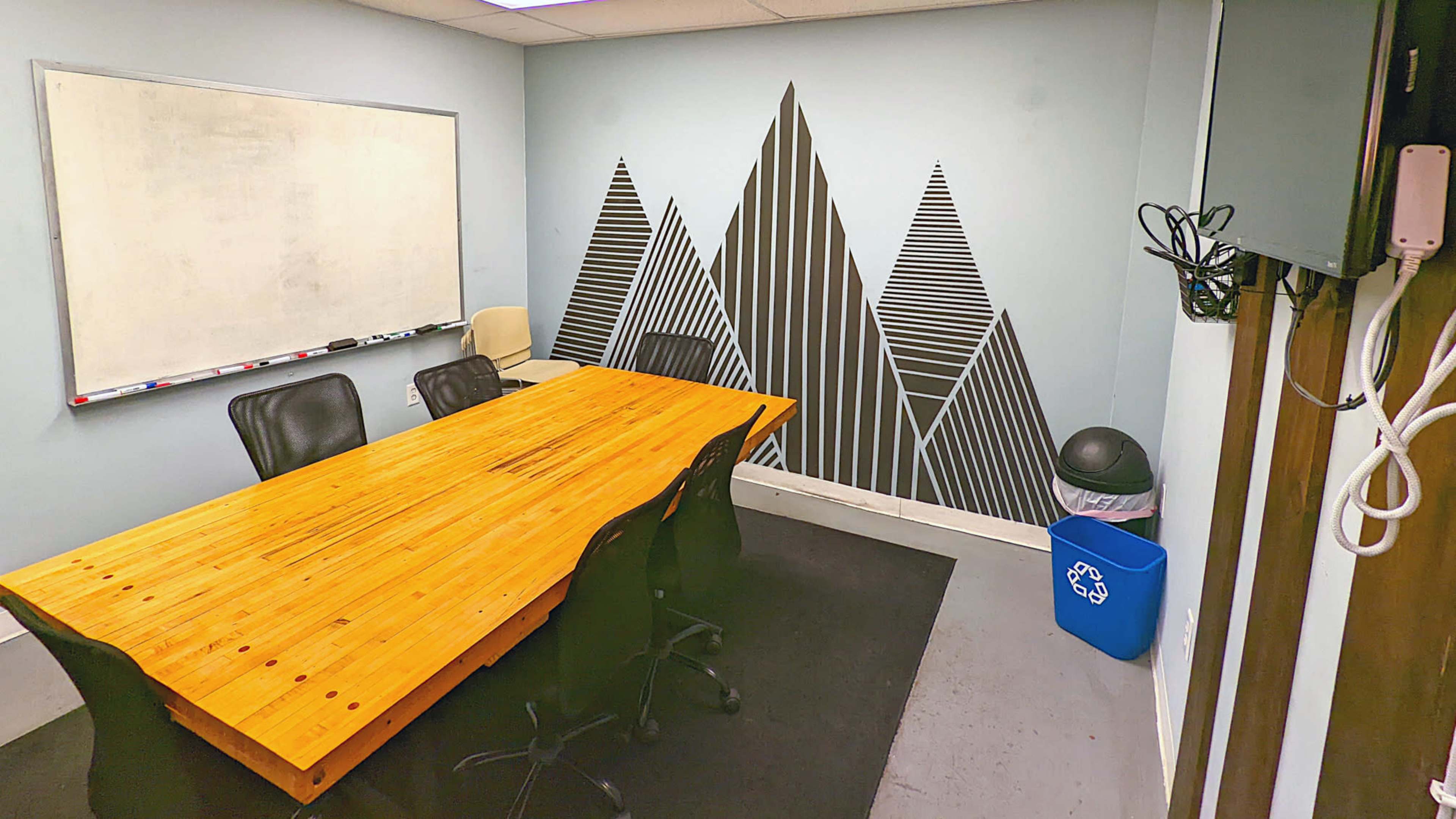 A conference room features a long wooden table surrounded by black chairs, with a whiteboard on one wall and a patterned mural of mountains on another wall.