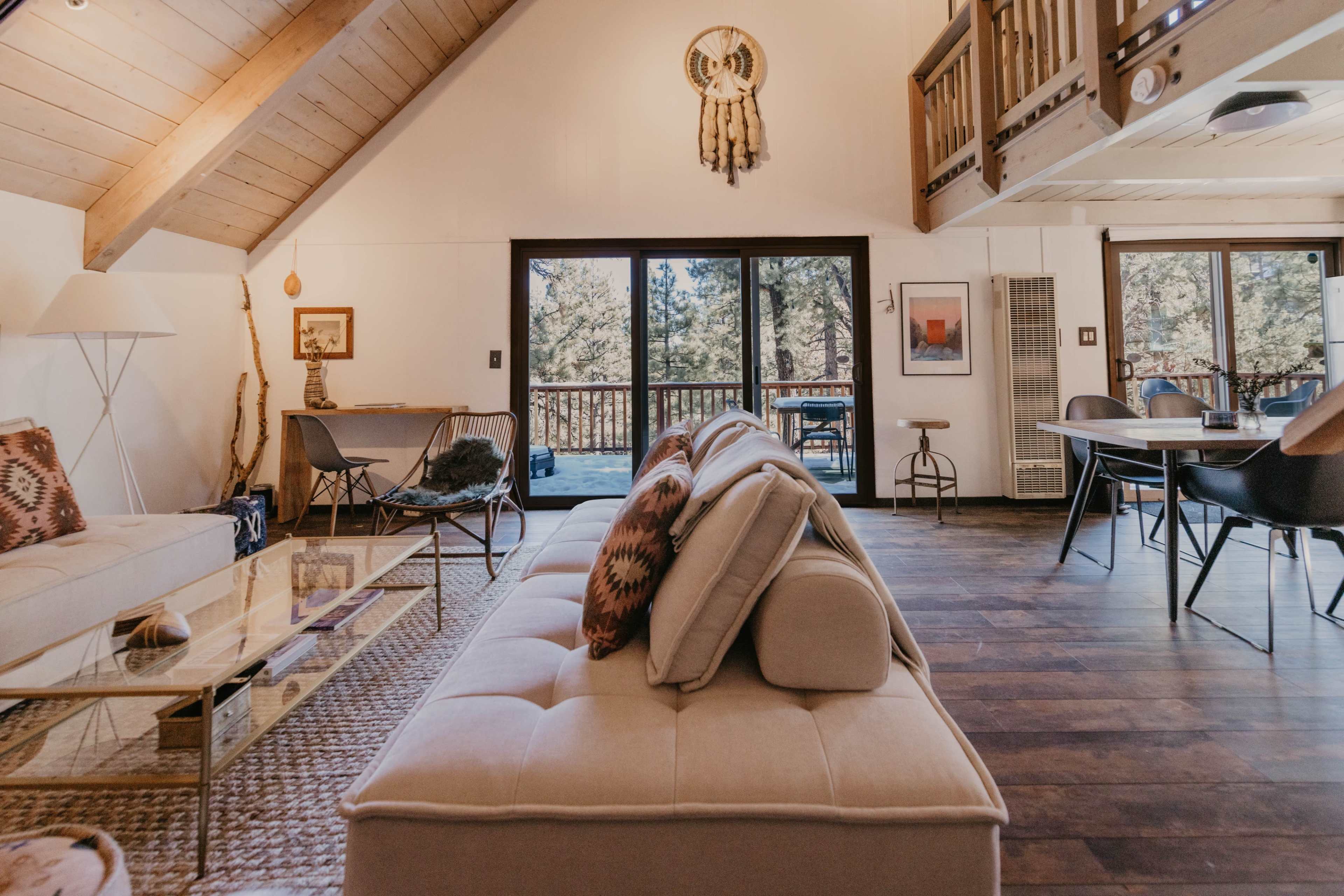A cozy living area with a light-colored sofa, a glass coffee table, and large windows overlooking a wooded view.