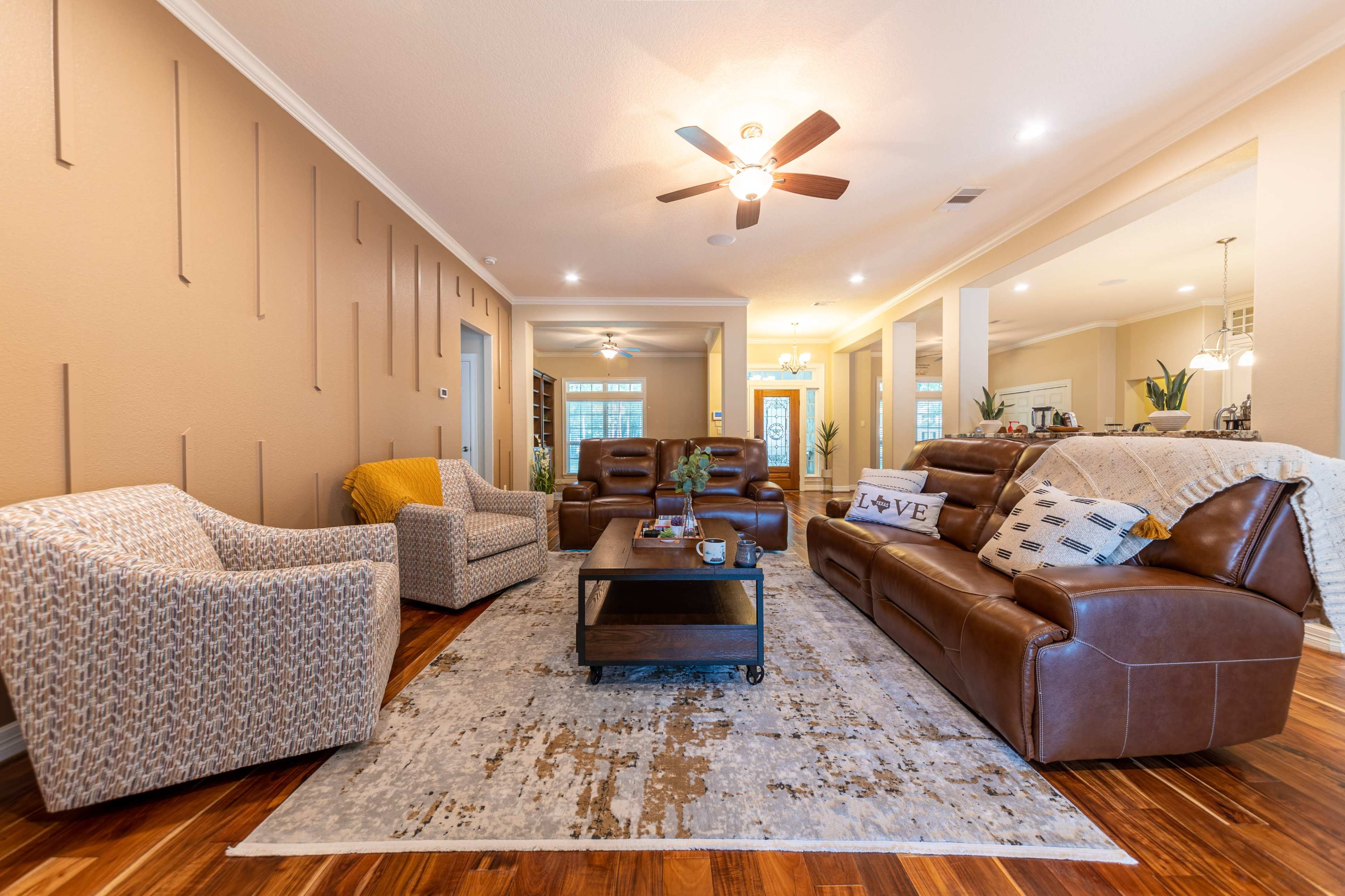 A spacious living room with two patterned armchairs, a brown leather sofa, a coffee table, and a large area rug, illuminated by a ceiling fan and overhead light.