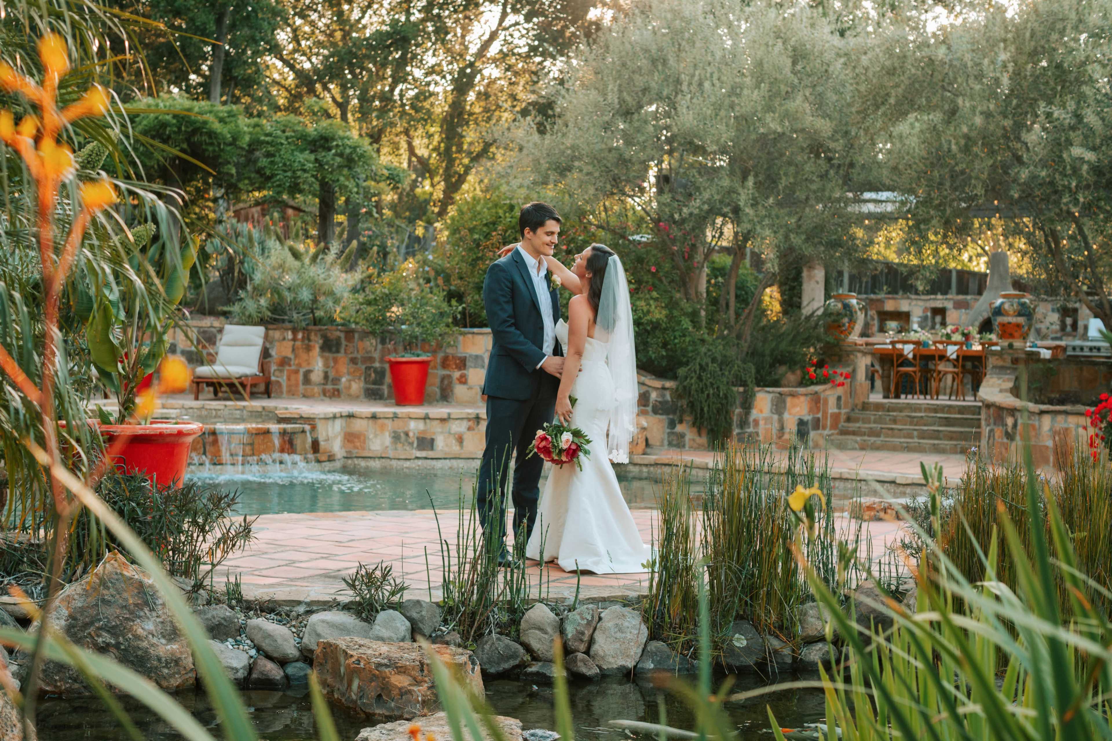 A bride and groom stand close together on a stone path beside a tranquil pond, surrounded by lush greenery and elegant wedding decor.