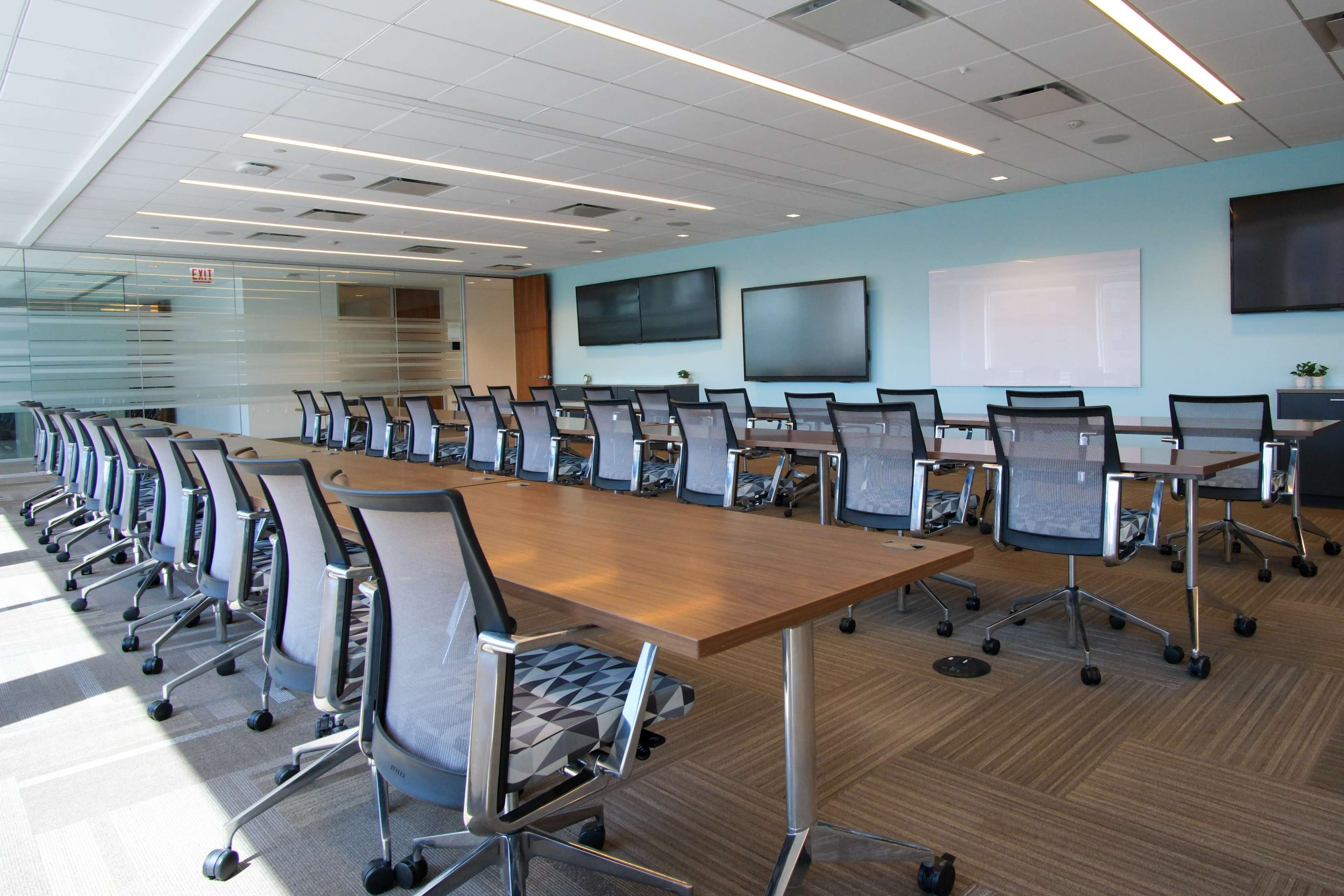 The image shows a modern conference room equipped with a long table surrounded by numerous ergonomic chairs, large screens on the walls, and bright overhead lighting.