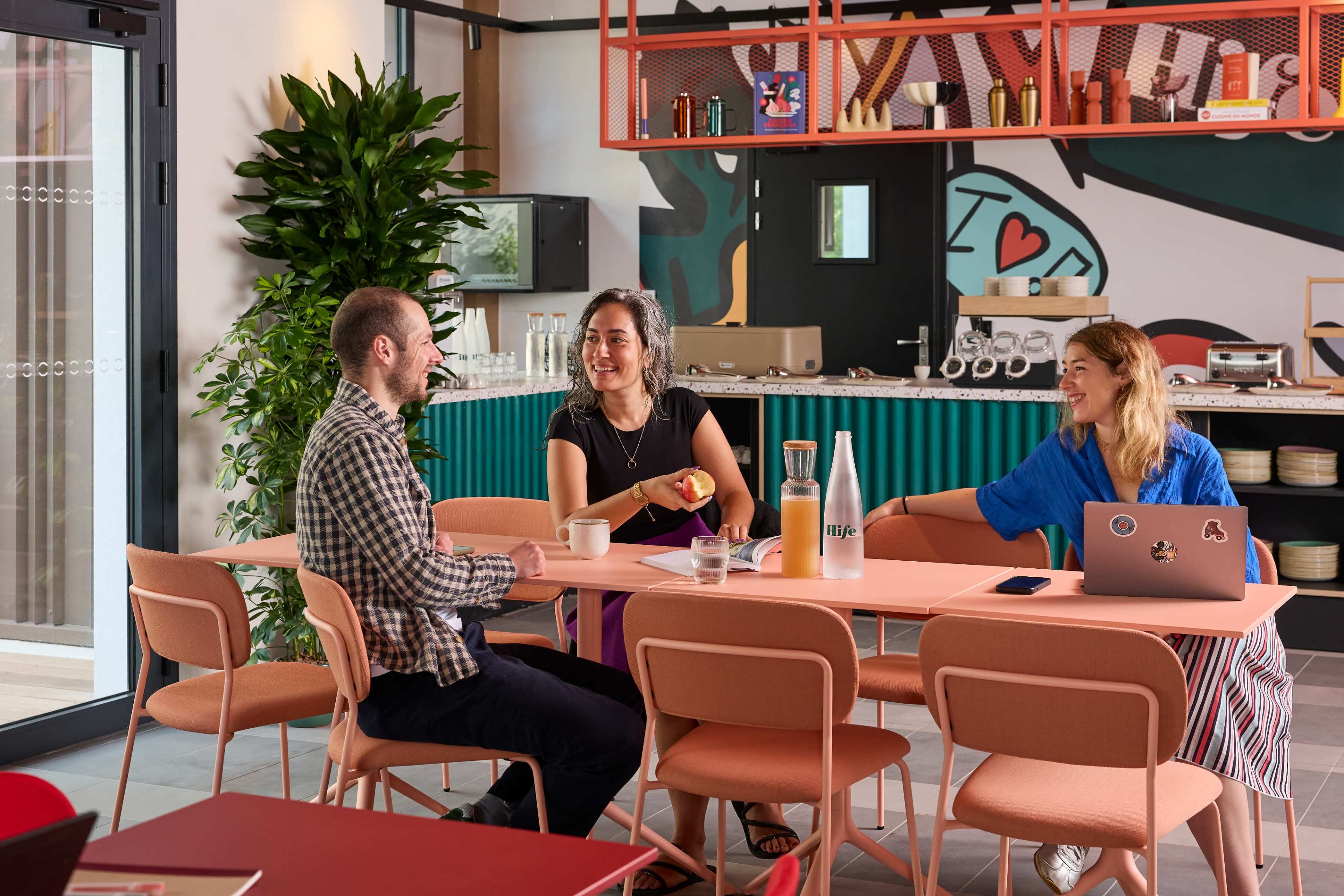 Three people are seated at a table in a modern, colorful workspace, engaged in conversation with drinks and a laptop on the table.