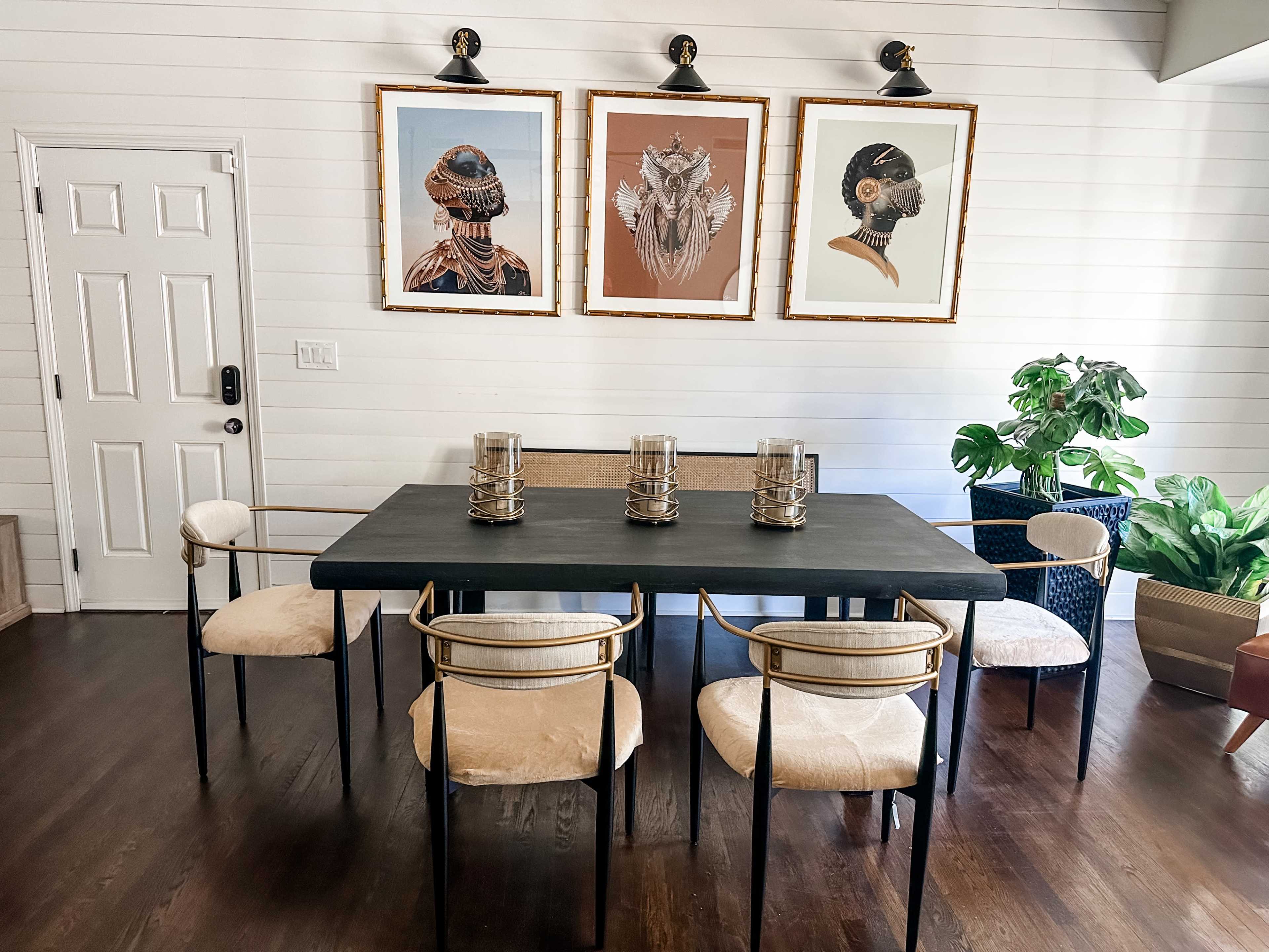 The image shows a dining area with a black table surrounded by six chairs, two of which have a light upholstery, and three framed artworks on the wall above.