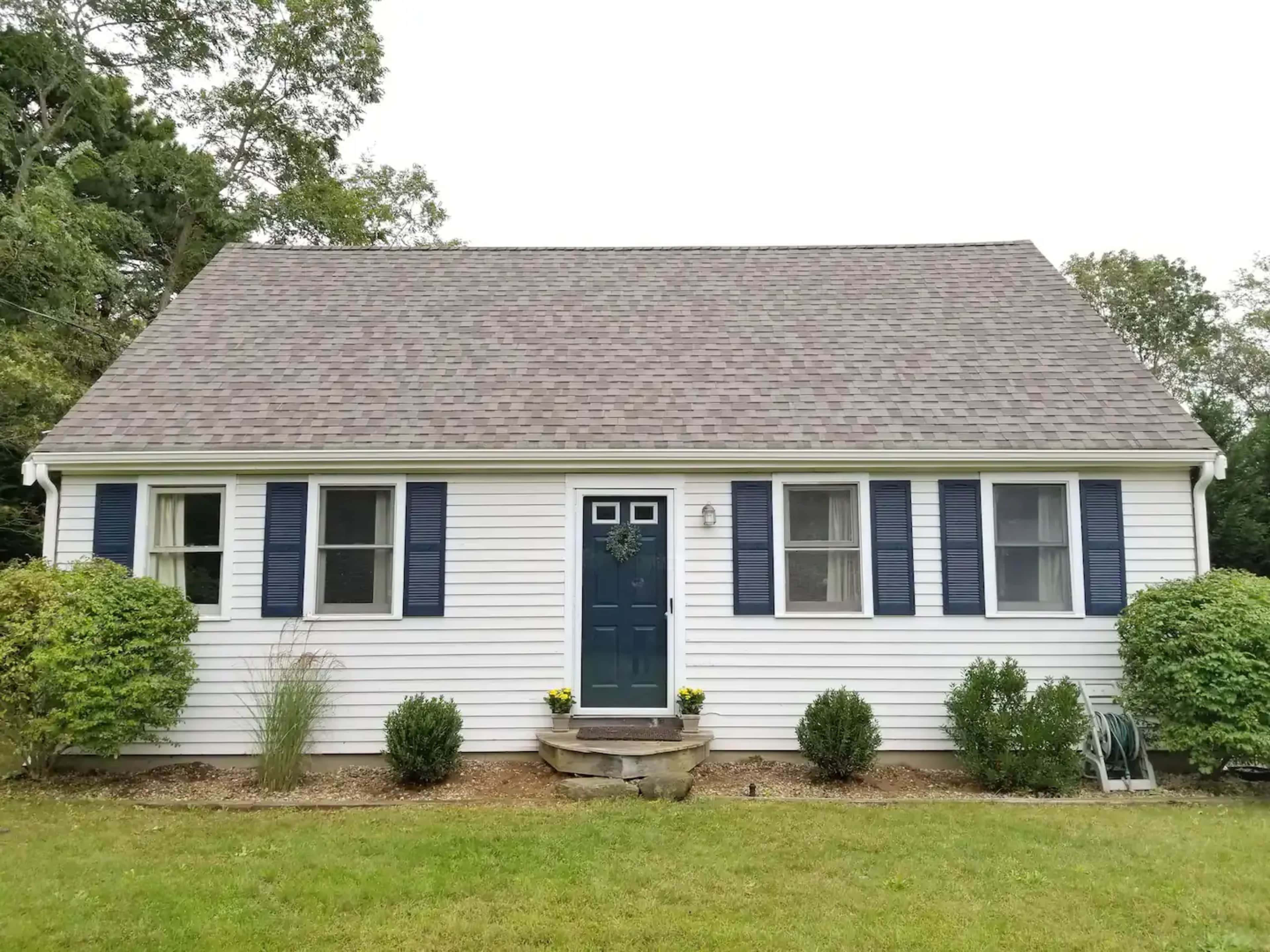 A single-story house features a blue front door, white siding, and dark blue shutters on the windows, surrounded by neatly trimmed shrubs.