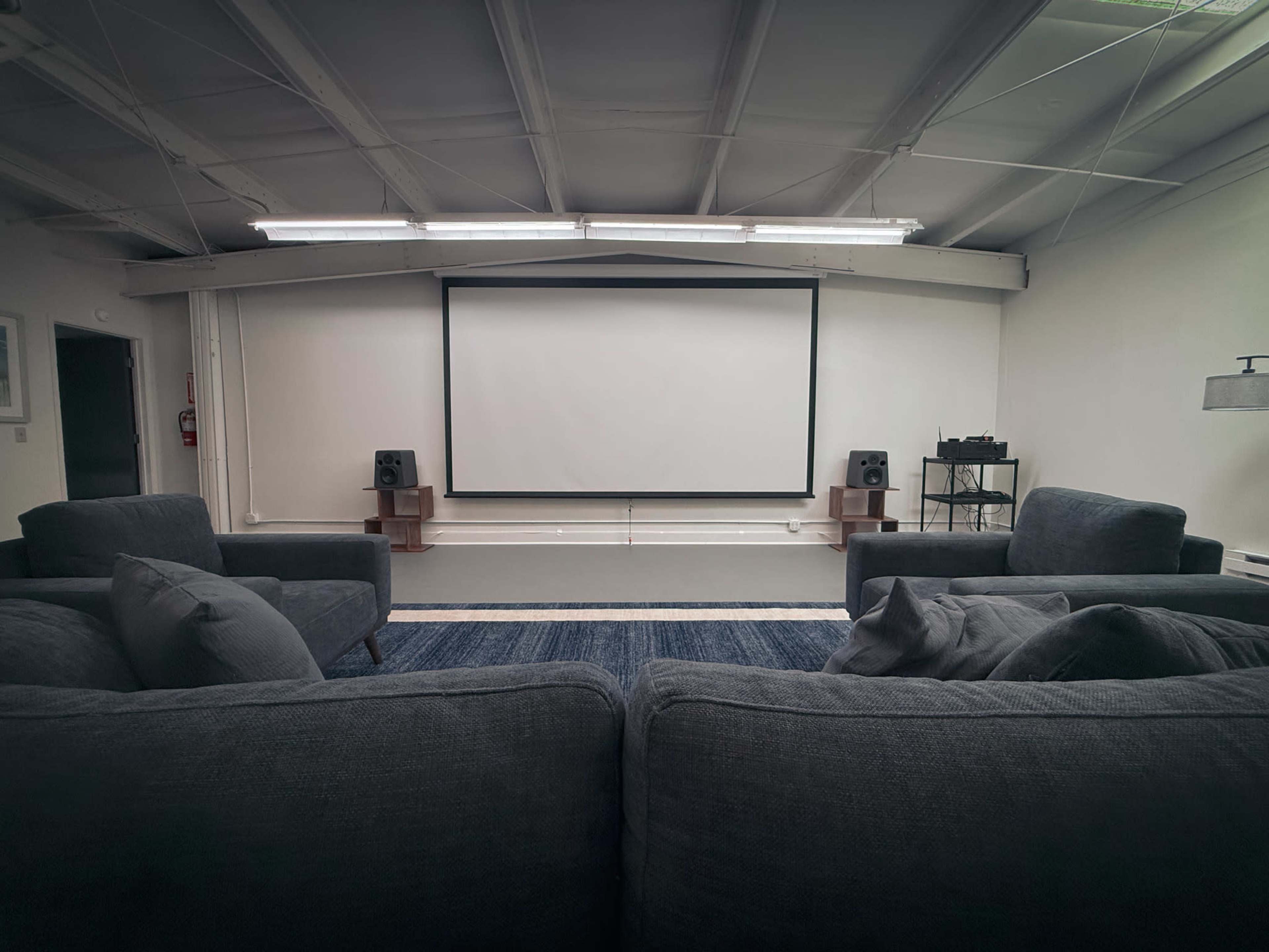 A well-lit room features a large projection screen at the front, flanked by two speakers and furnished with several dark sofas arranged in a semi-circle.