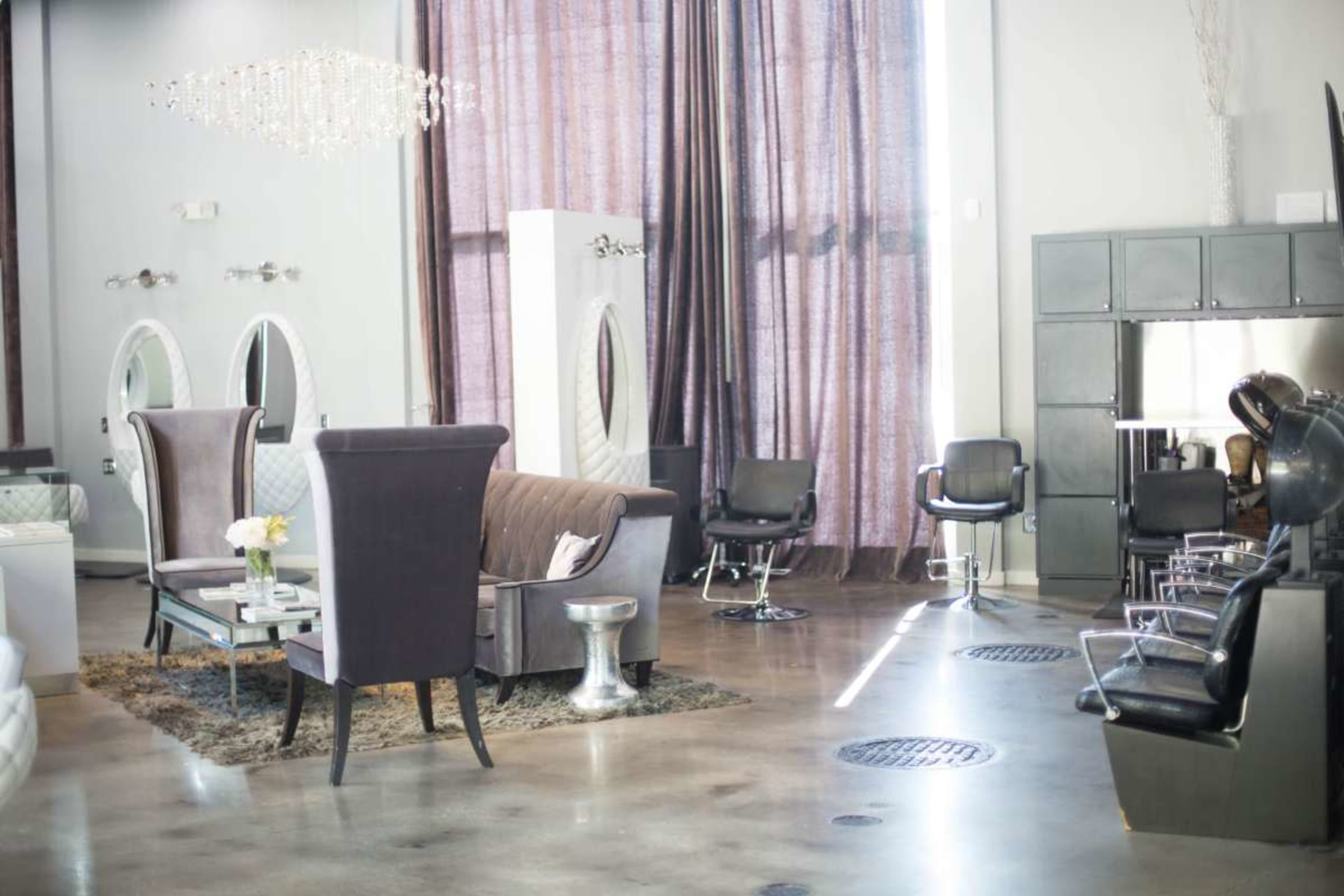 The image shows a modern salon interior featuring a seating area with a sofa and chairs, large windows with curtains, and styling stations with salon chairs.
