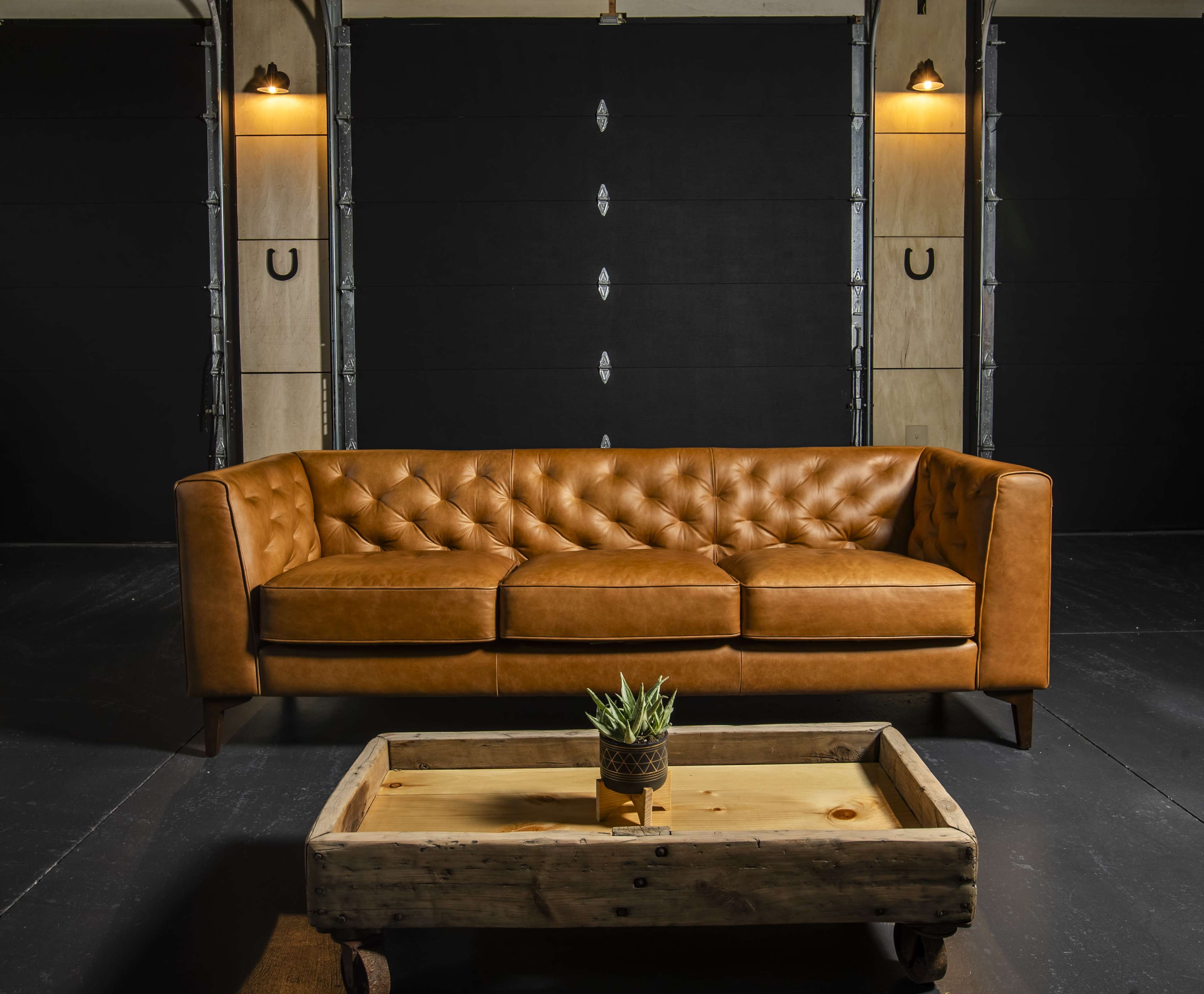 A tufted leather sofa in a warm brown color sits in front of a rustic coffee table on a dark wooden floor, with industrial-style lighting and a backdrop featuring a textured wall.