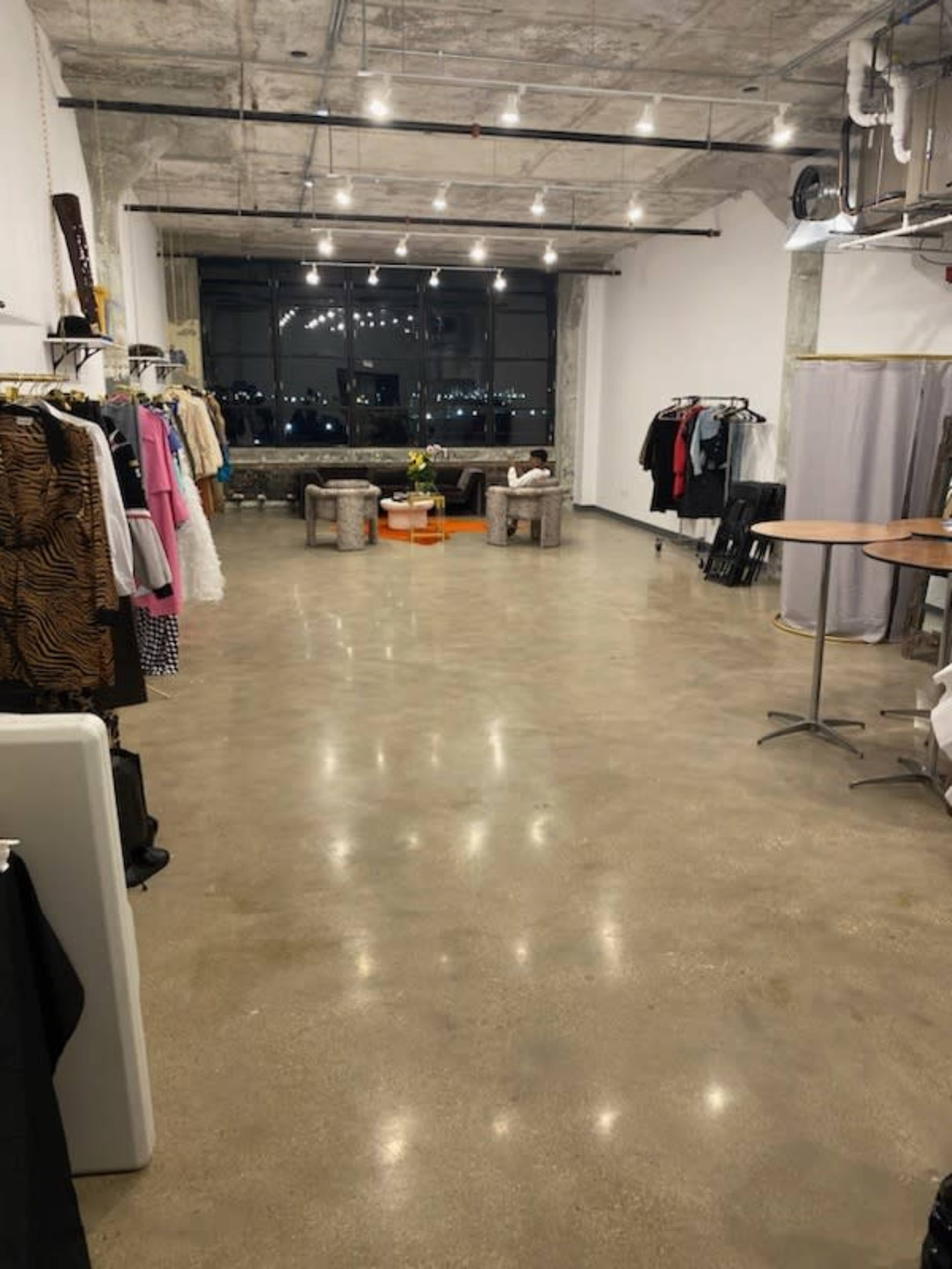 The image shows an empty, well-lit studio space with clothing racks on the sides and a seating area near a large window.