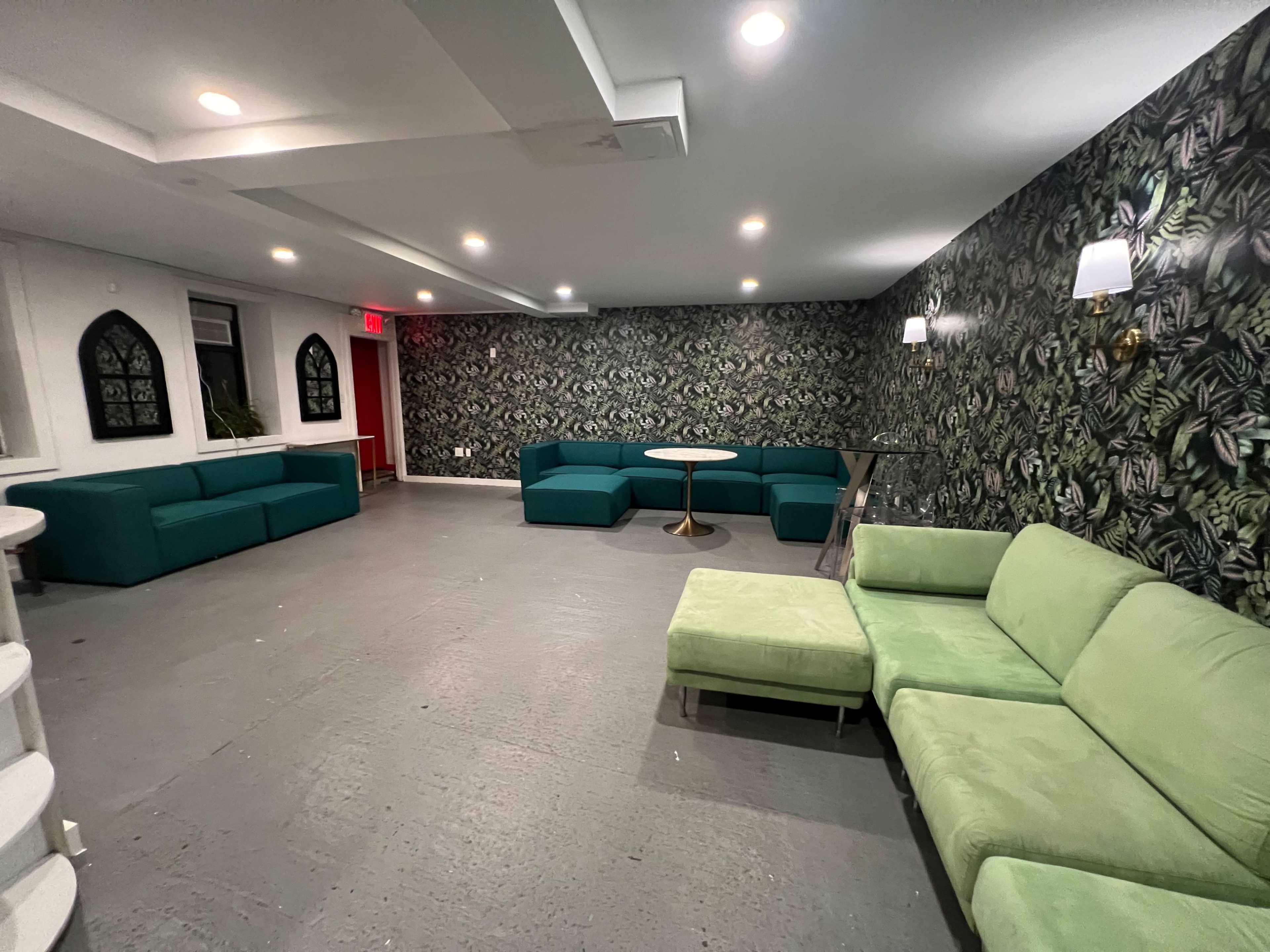 A spacious lounge area features a green sofa set and a round table against a wallpapered wall with a floral design.