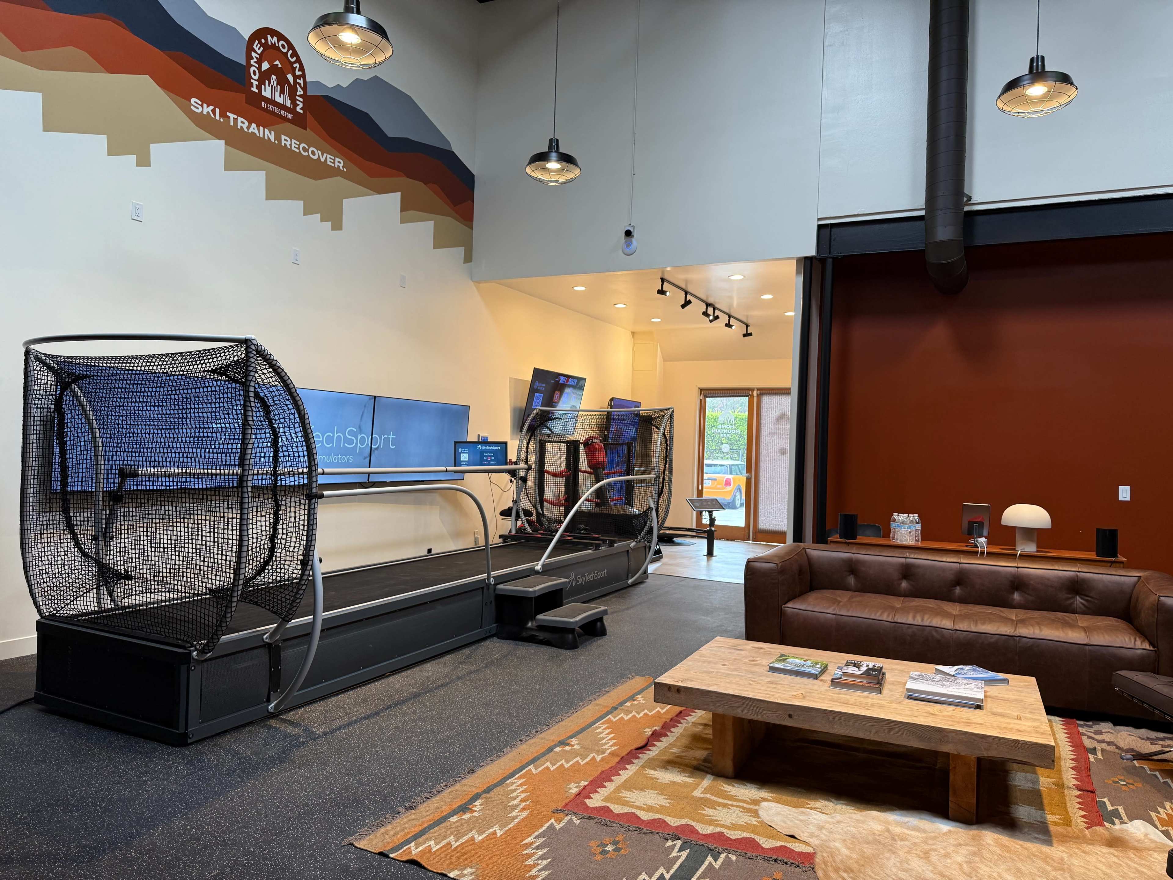 The image shows a modern training facility featuring a simulated ski treadmill with a netted enclosure, a reception area with screens, and a seating area with a leather couch and coffee table.