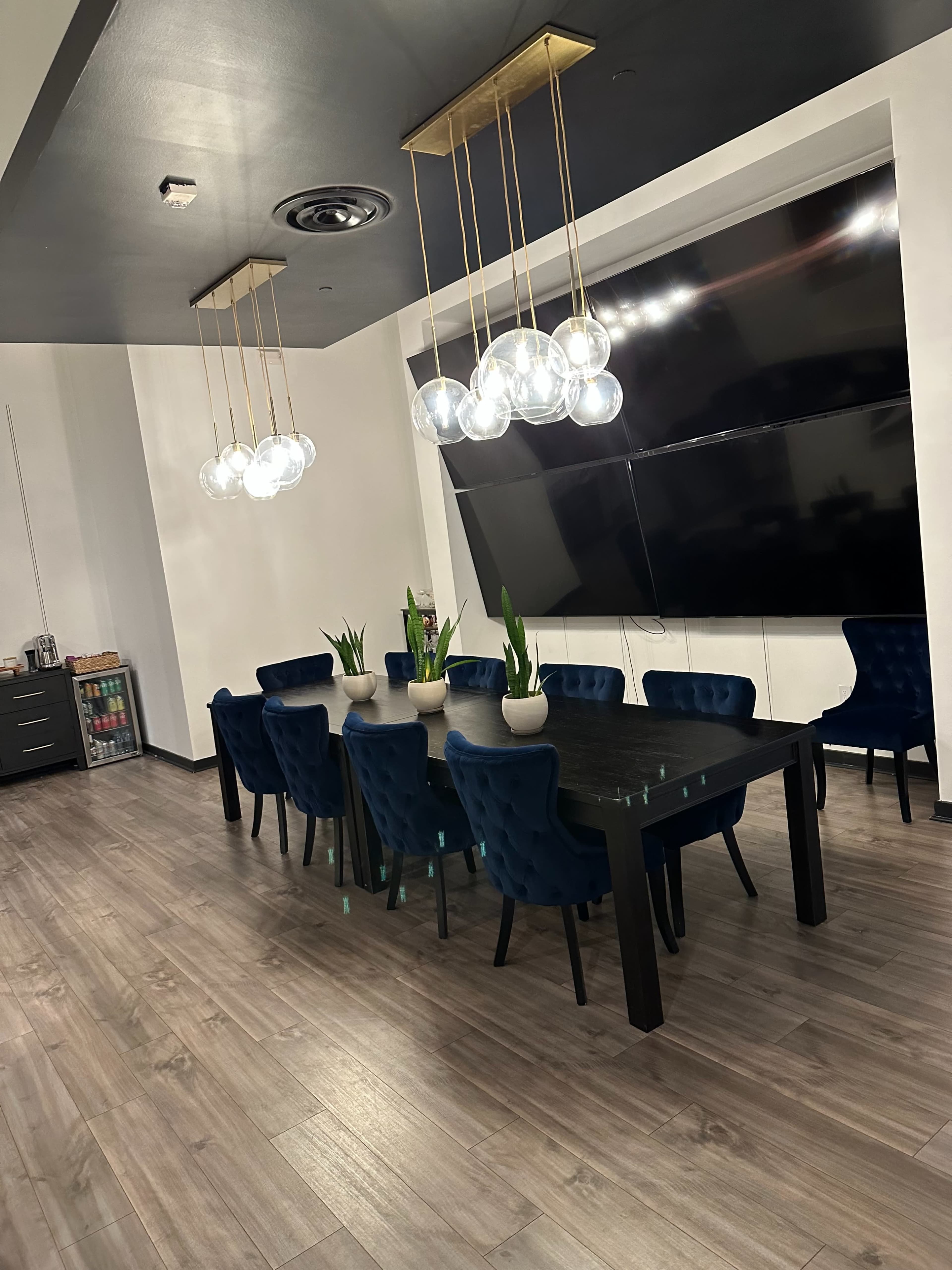A modern dining room features a long black table surrounded by blue upholstered chairs, with pendant lights hanging above and a large TV mounted on the wall.