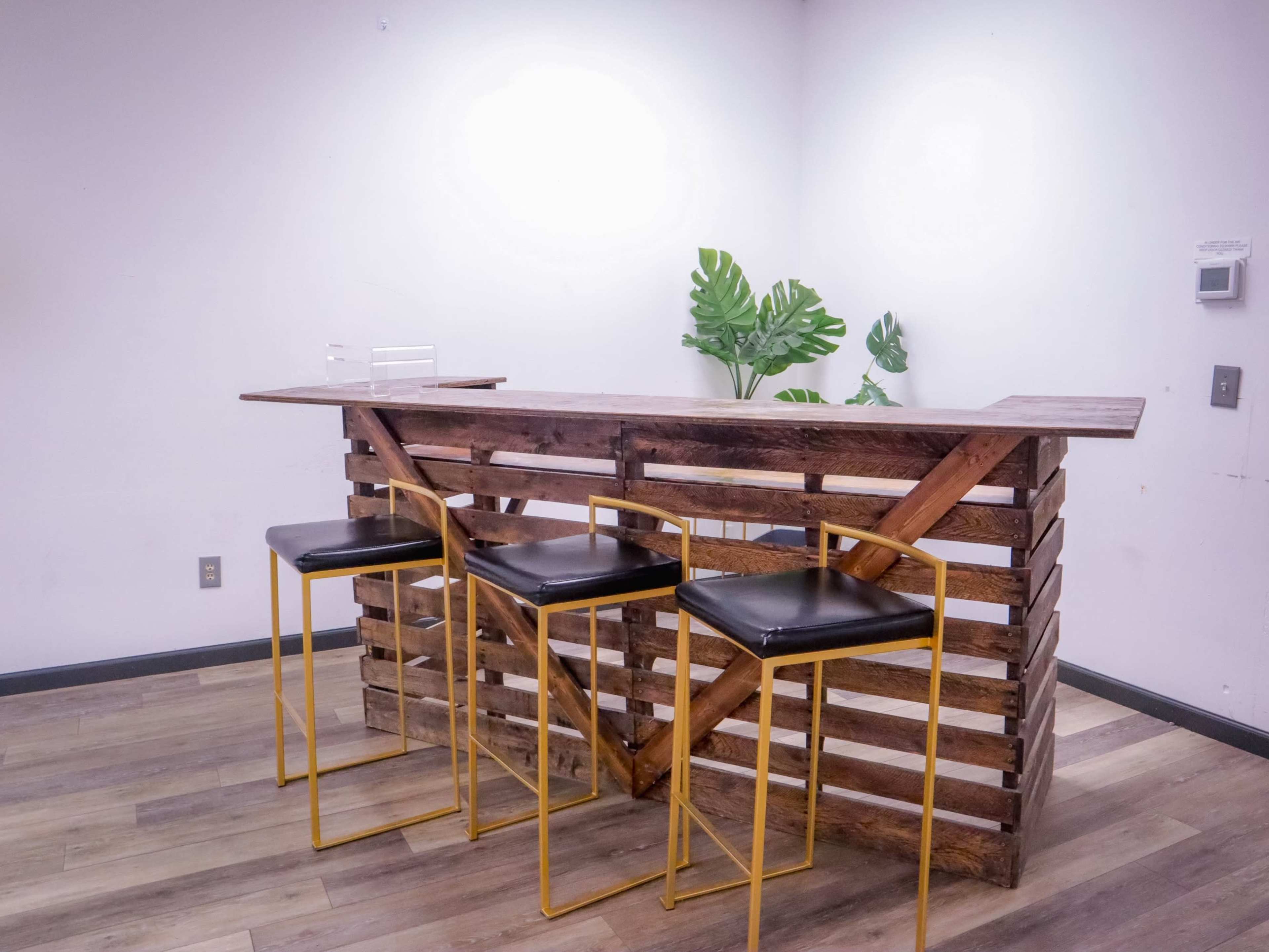 A wooden bar with a slatted design and three black stools stands against a plain wall, complemented by a nearby plant.