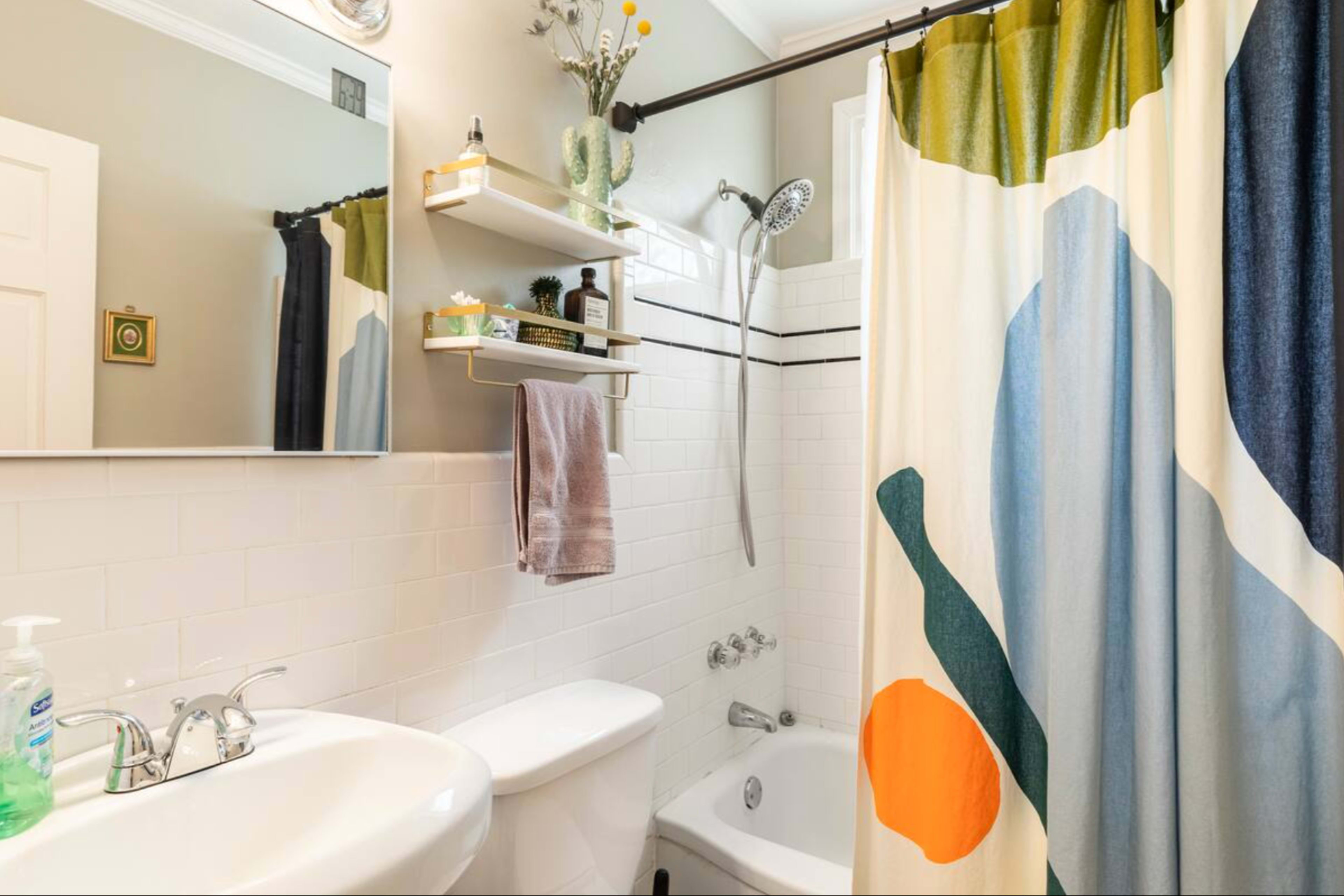 A small bathroom features a shower with a colorful shower curtain, a white sink and toilet, and shelves with toiletries.