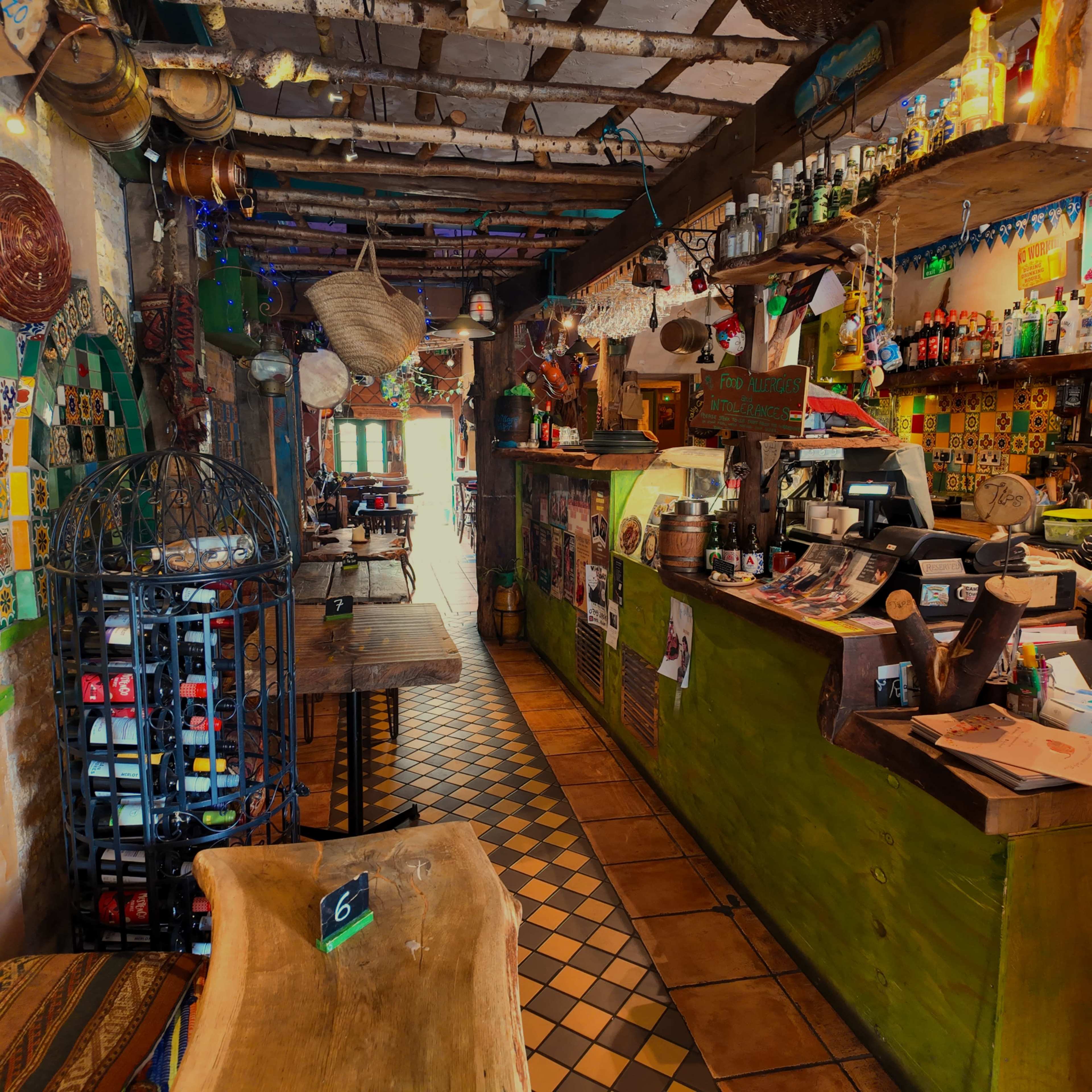 The image shows a vibrant, eclectic restaurant interior with rustic wooden tables, colorful tile work, and a bar filled with various bottles.