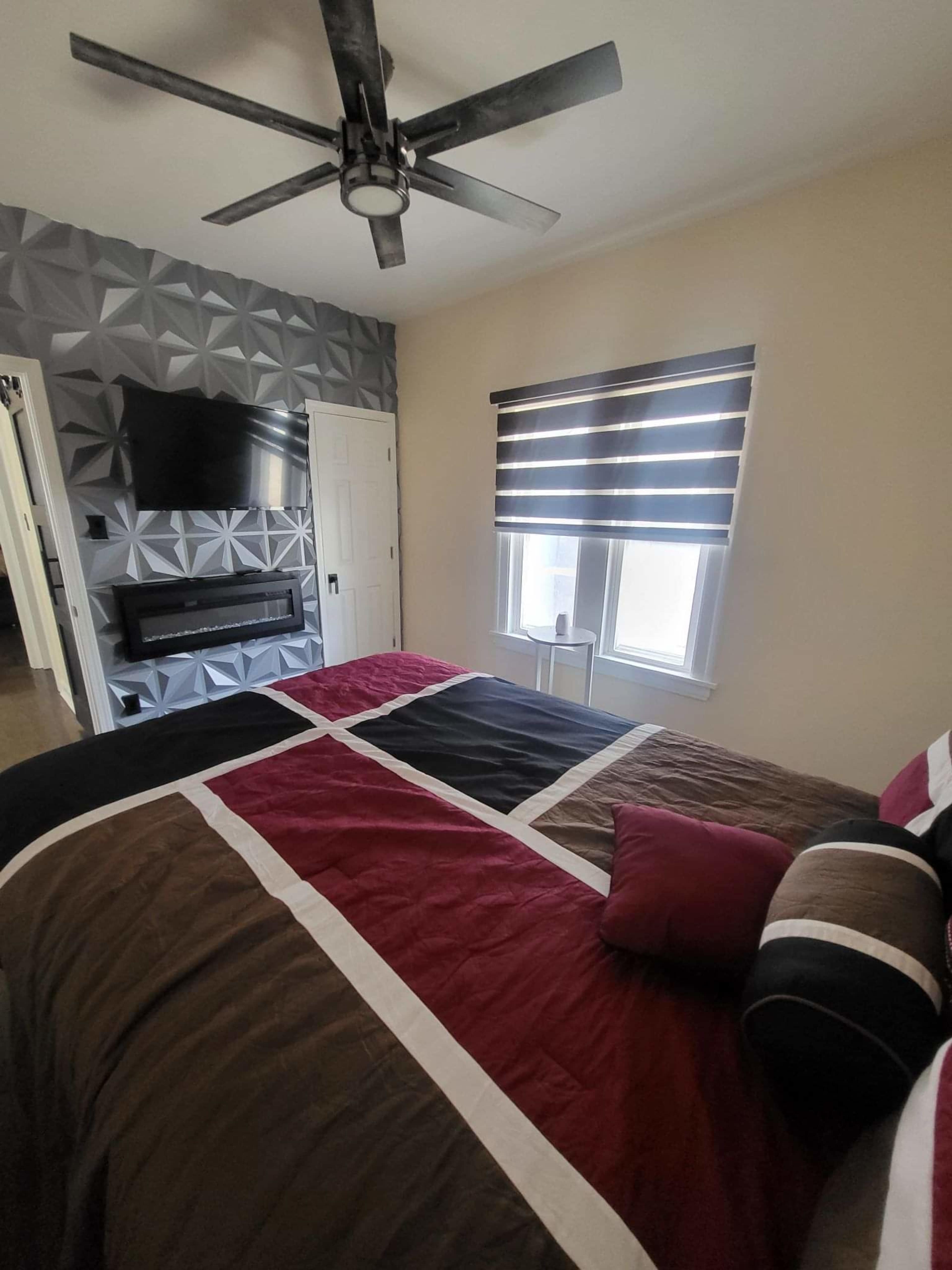 The image shows a modern bedroom featuring a patterned accent wall, a bed with a multi-colored duvet, a wall-mounted TV, and black-out roller shades on the windows.