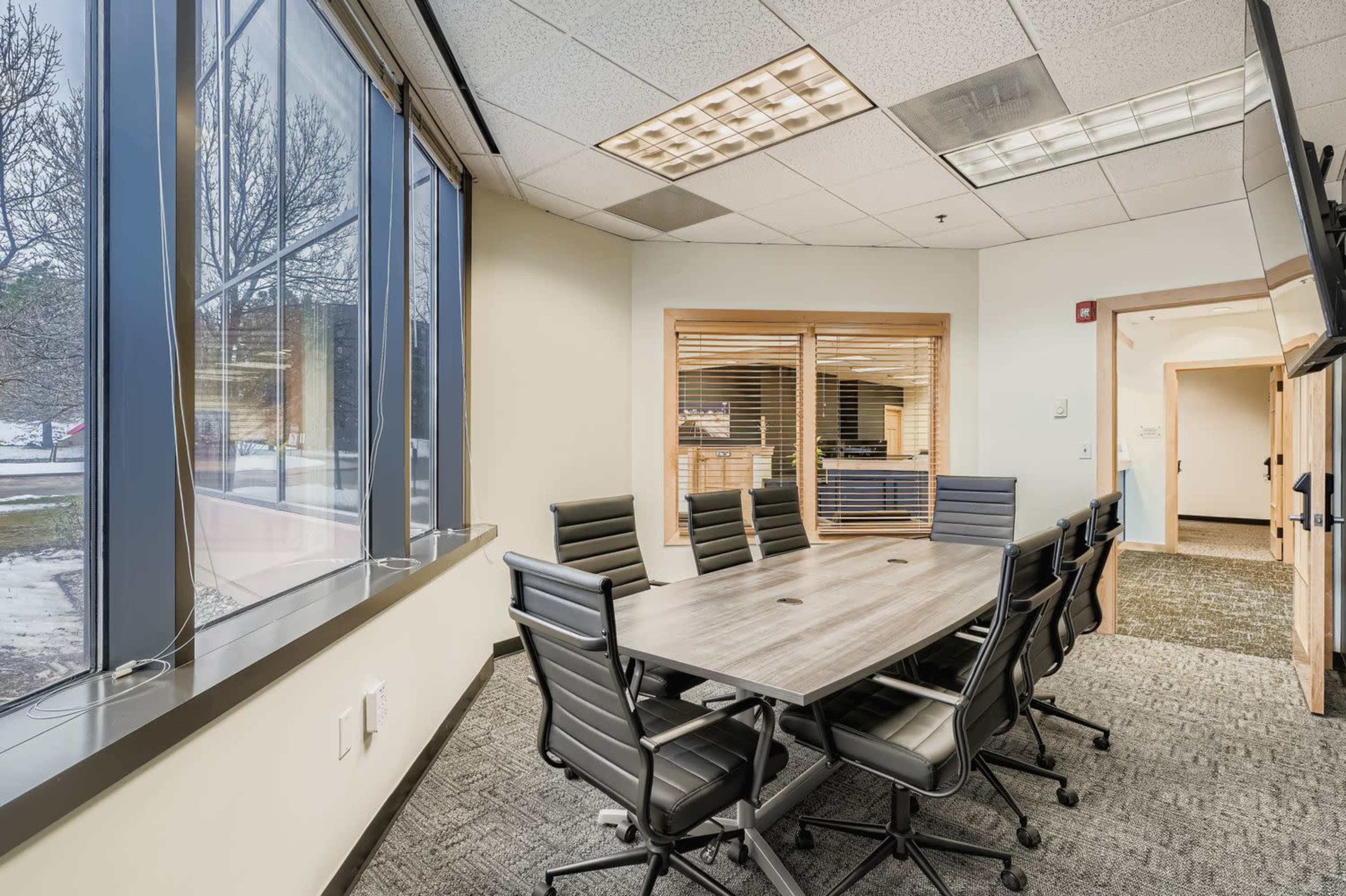 The image shows a modern conference room featuring a large table surrounded by black ergonomic chairs, with windows providing natural light.