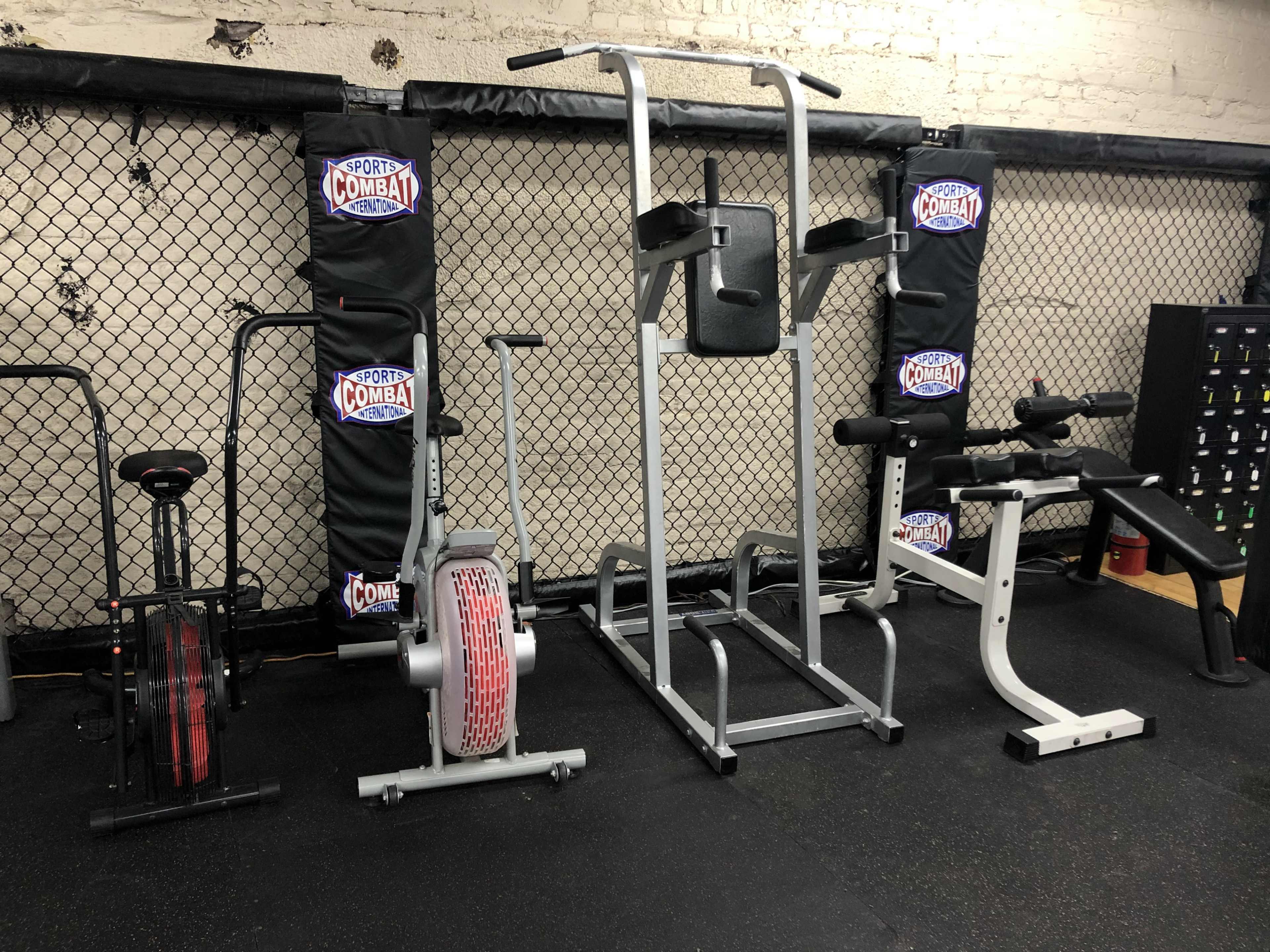 A gym area with various exercise equipment, including a rowing machine, a pull-up station, and a weight bench, all set against a chain-link fence with branding.