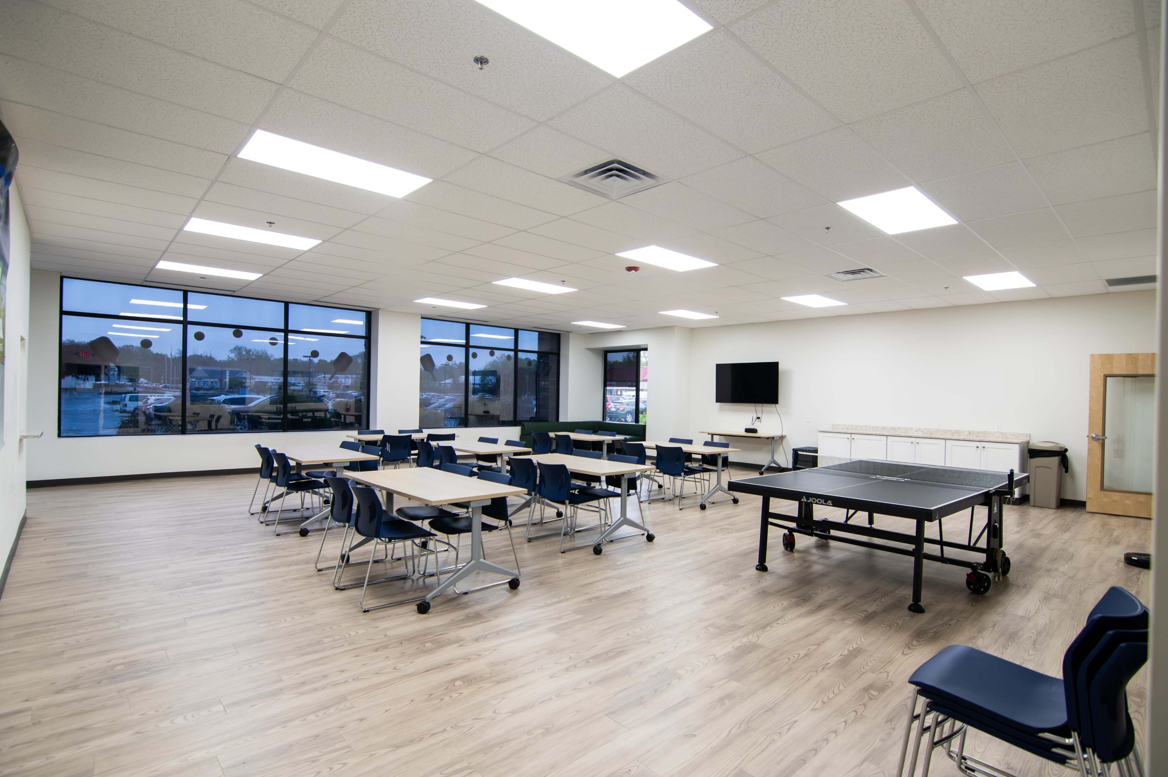 The image shows a spacious classroom with multiple tables and chairs arranged, a ping pong table, and large windows letting in natural light.