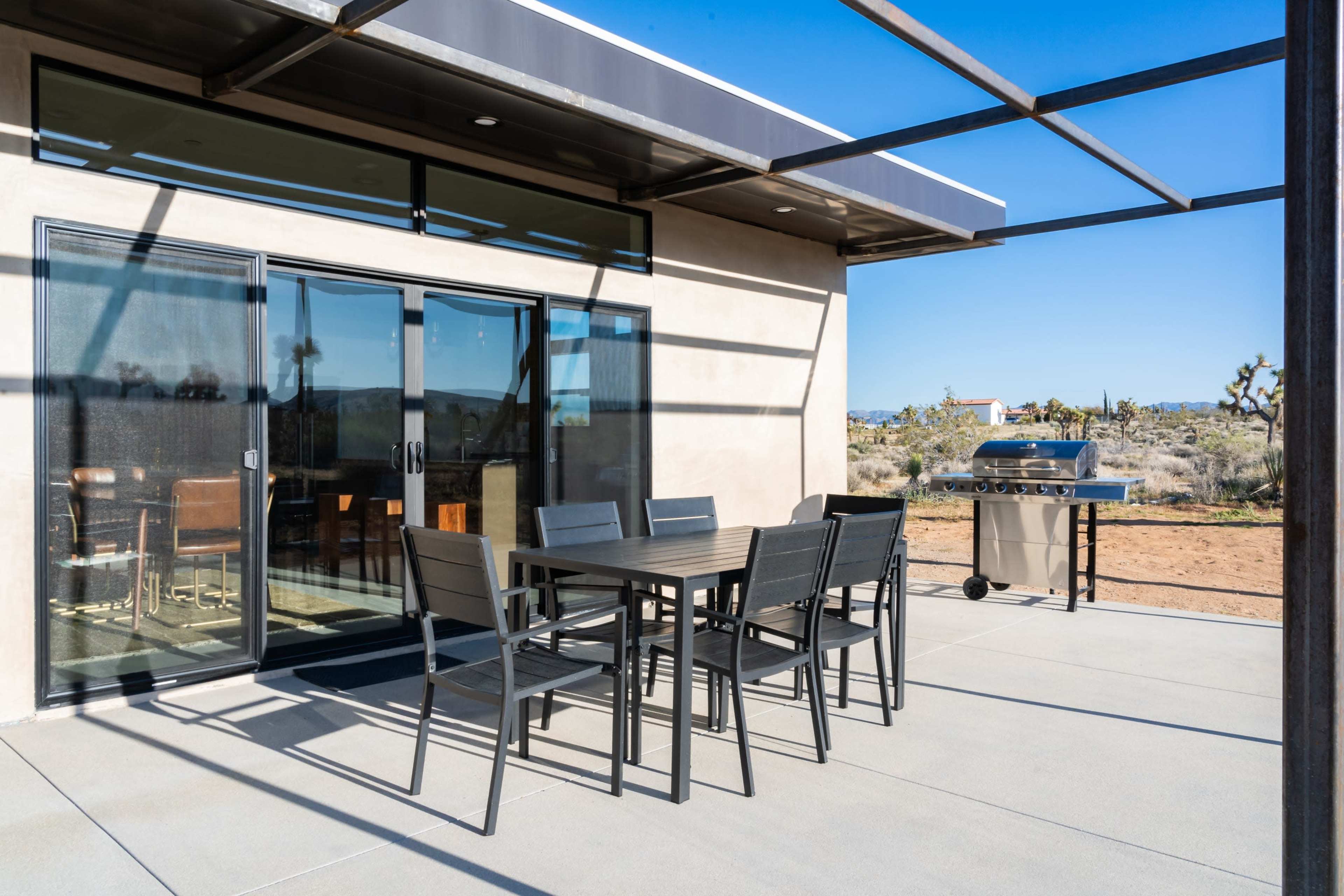 The image shows a modern outdoor patio area with a dining table and chairs adjacent to a barbecue grill, set against a desert landscape.