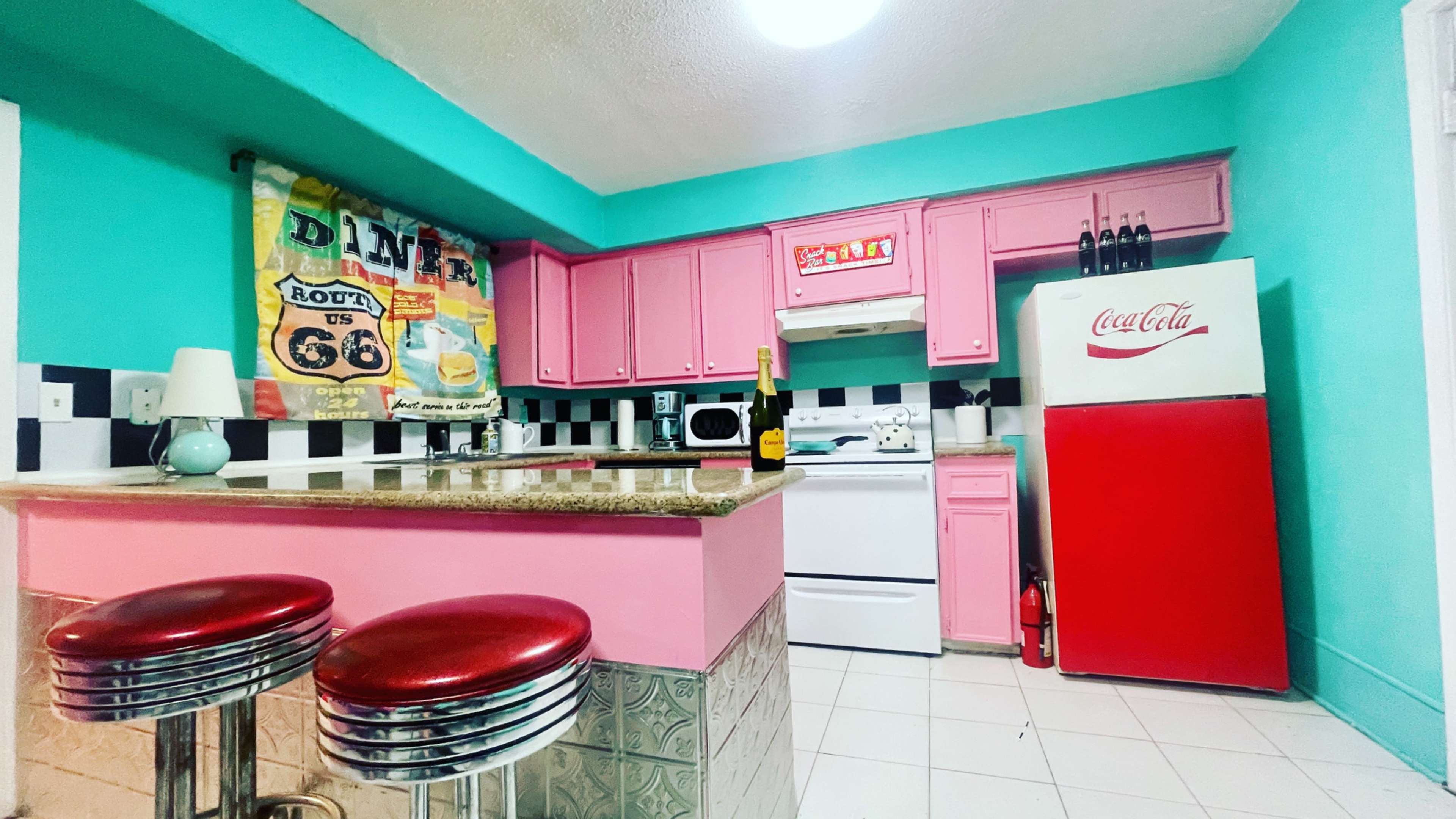 A brightly colored kitchen featuring pink cabinets, a red Coca-Cola fridge, and a bar area with chrome stools.