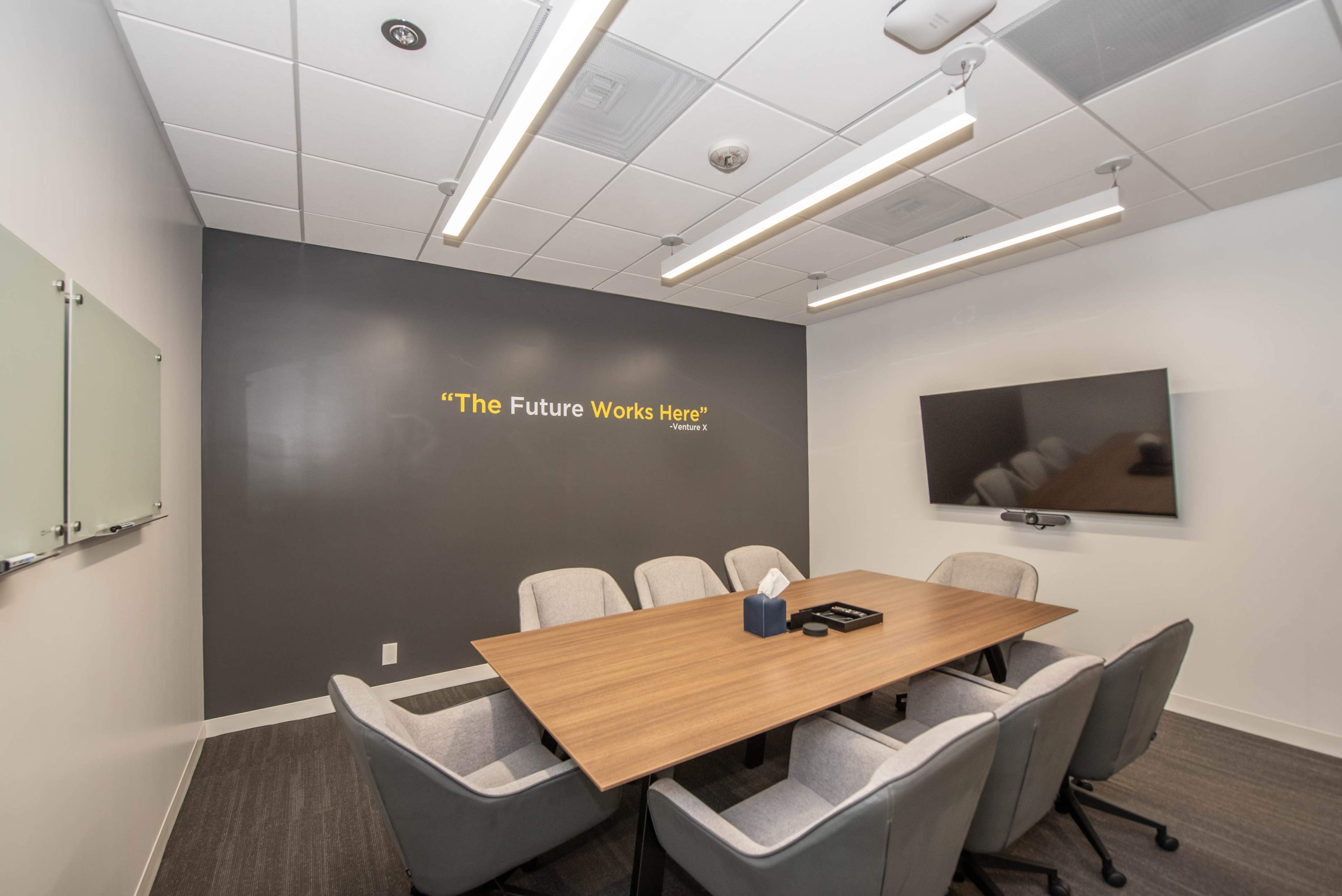 A conference room features a large wooden table surrounded by six gray chairs, with a wall displaying the phrase "The Future Works Here" and a mounted TV.