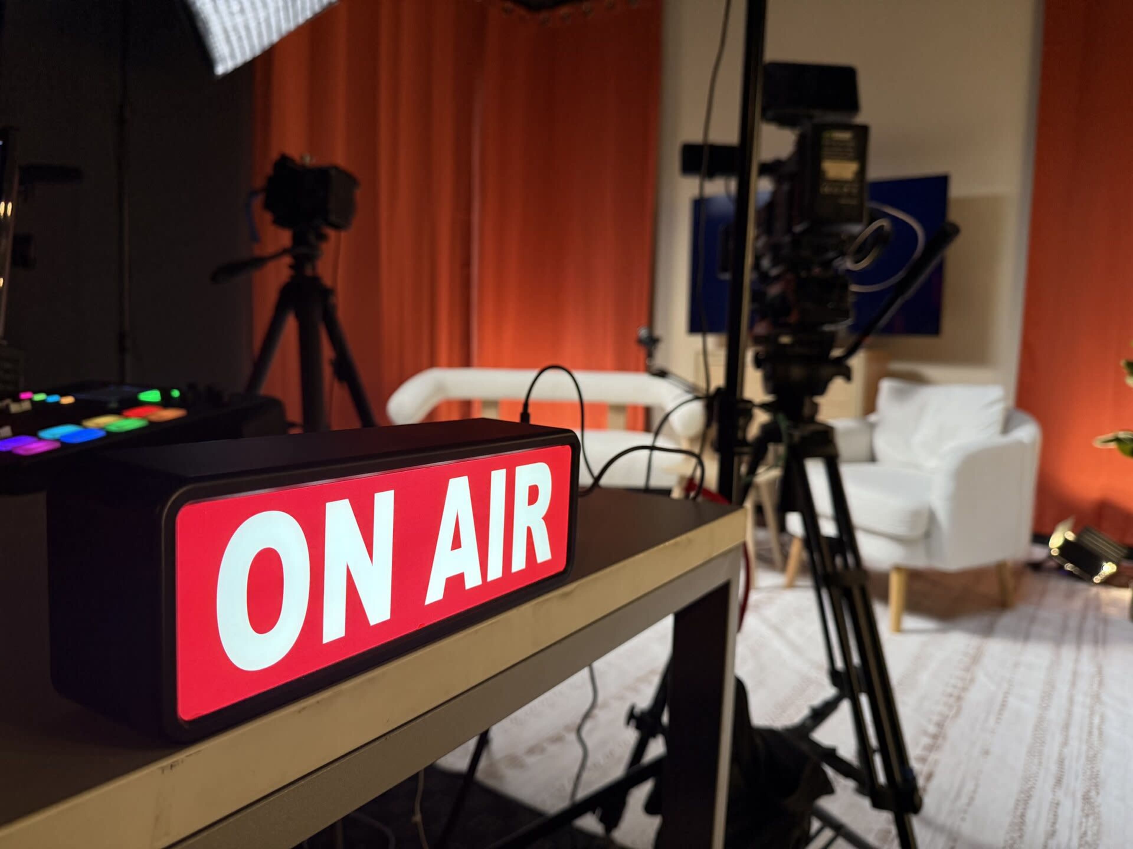 A bright red "ON AIR" sign is illuminated in a studio setting with cameras and a white chair in the background.