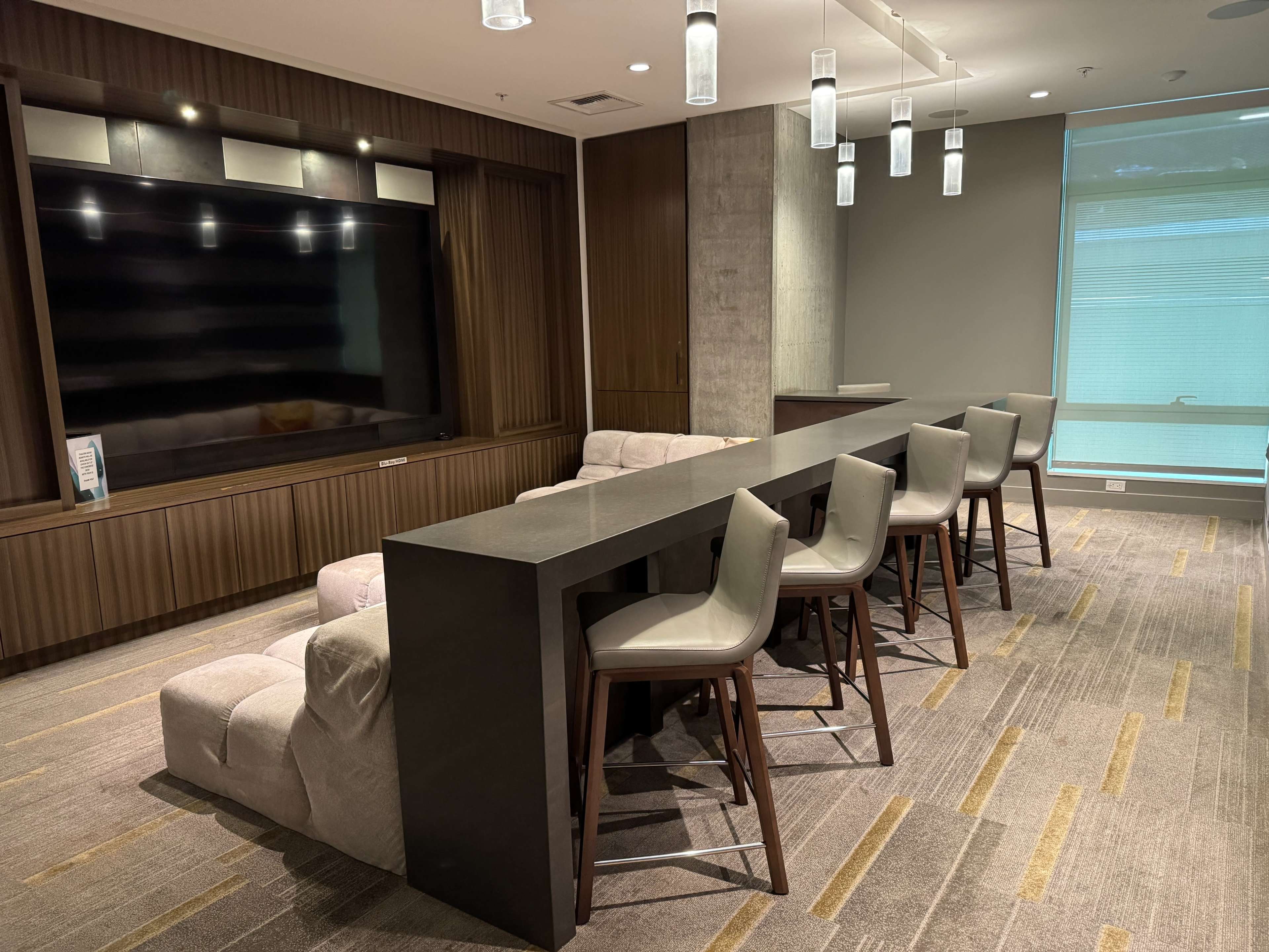 The image shows a modern lounge area with a large television, a gray sectional sofa, and bar-style seating along a dark counter.