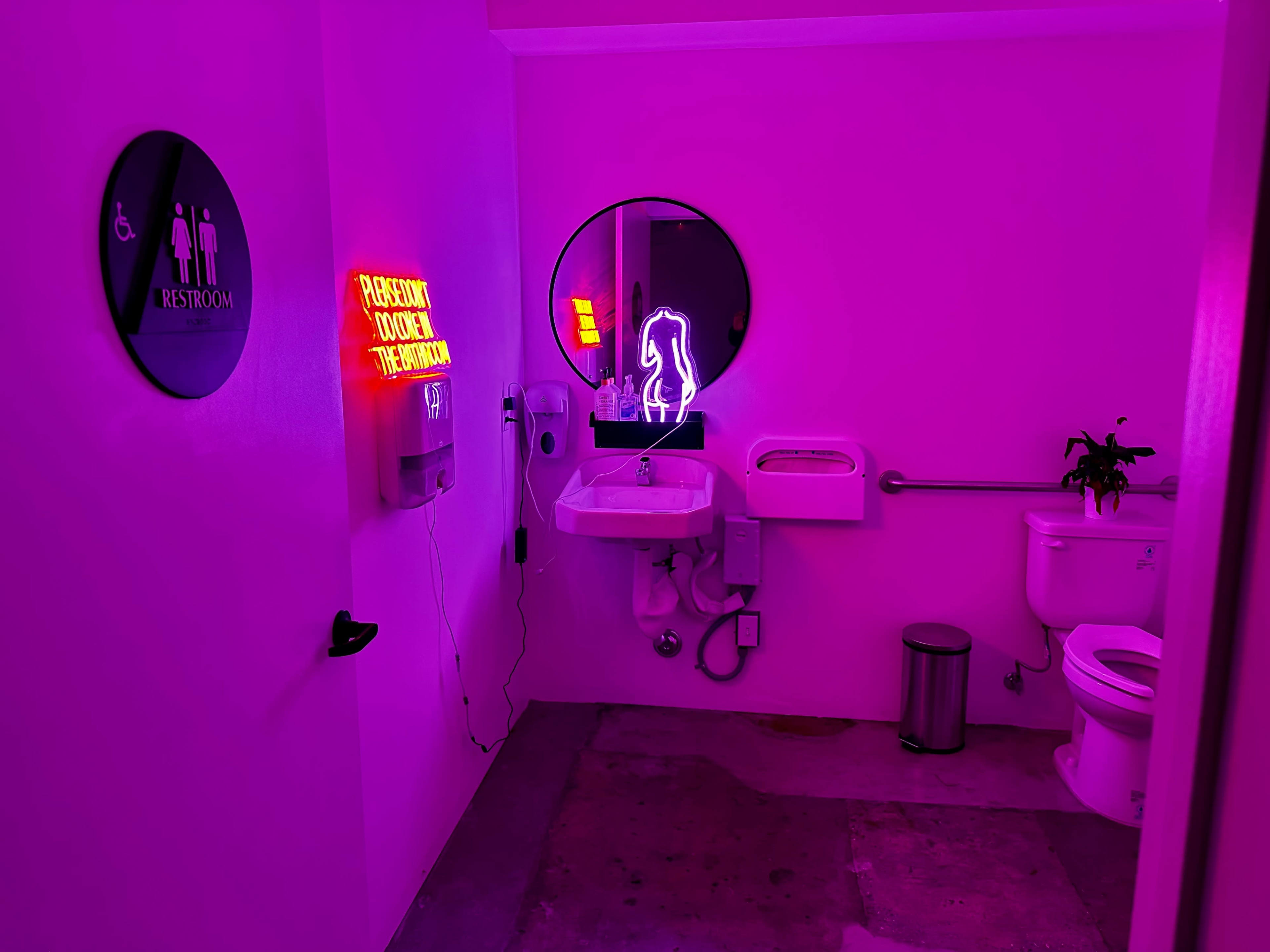 A brightly lit restroom featuring pink walls, a circular mirror, a sink, and a toilet, with neon signs displaying a horse and a message about restroom access.