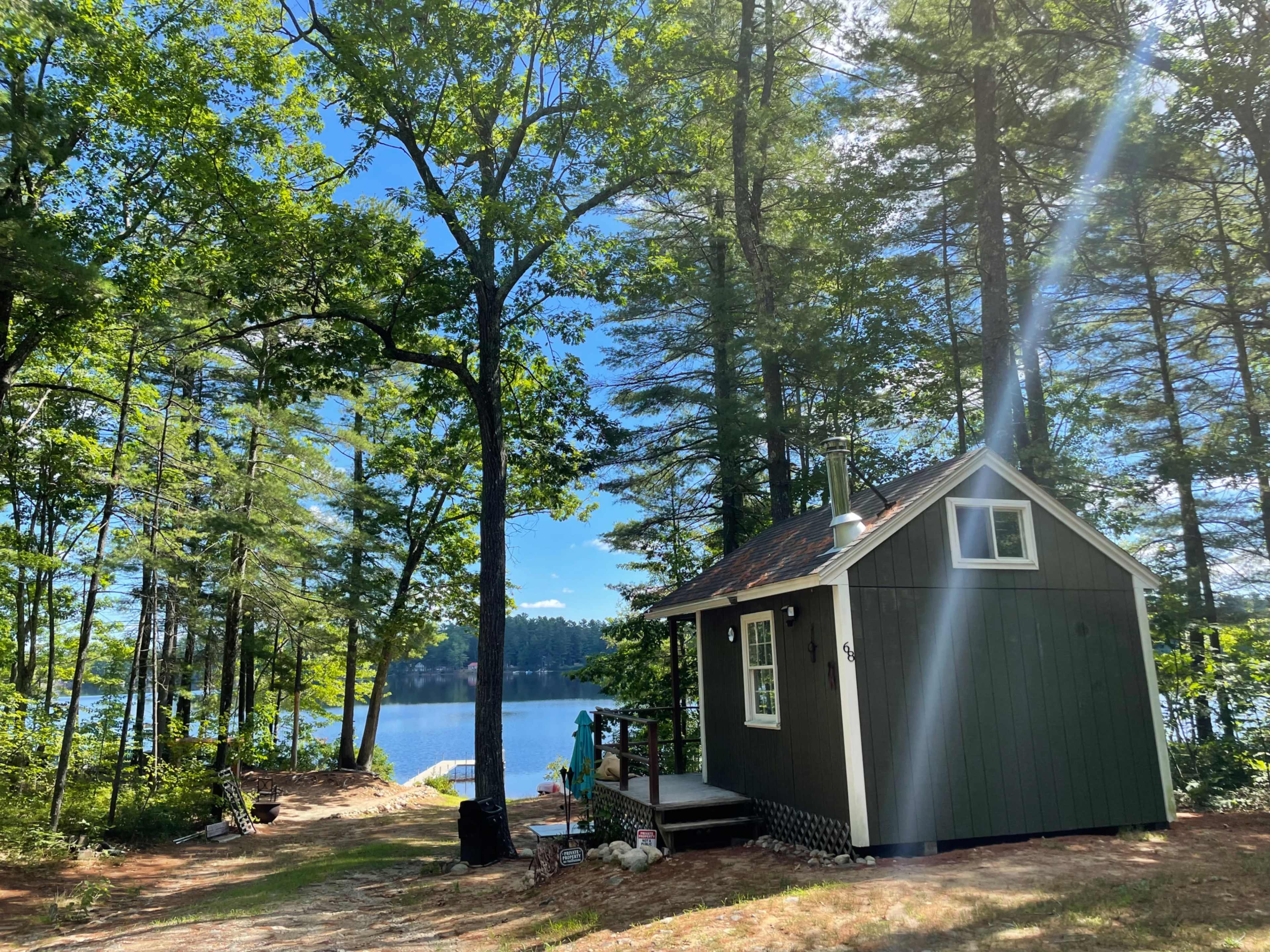 A small green cabin is situated near a lake, surrounded by tall trees and clear blue skies.