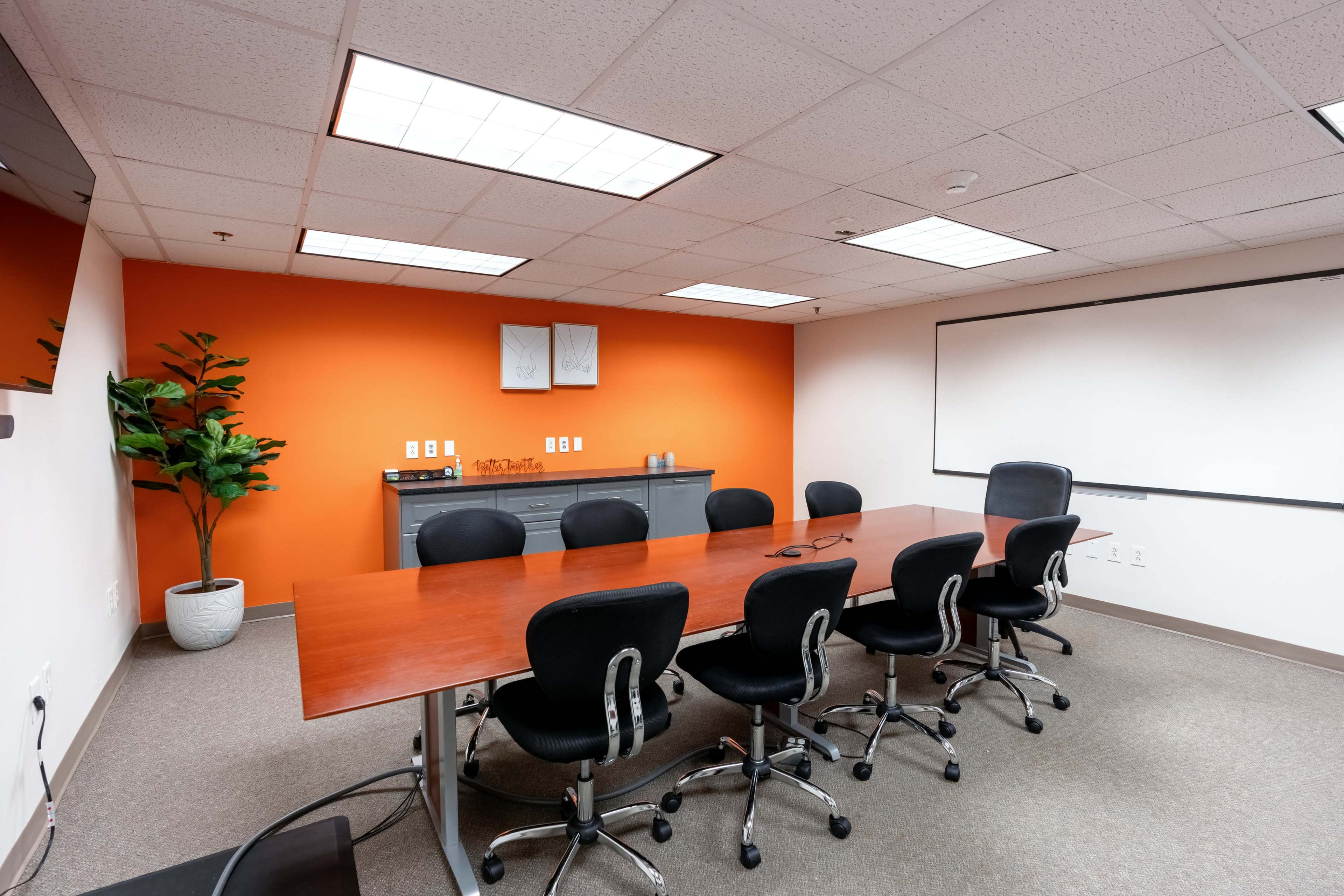 The image shows a meeting room with a large wooden table surrounded by black chairs, an orange accent wall, and a whiteboard.
