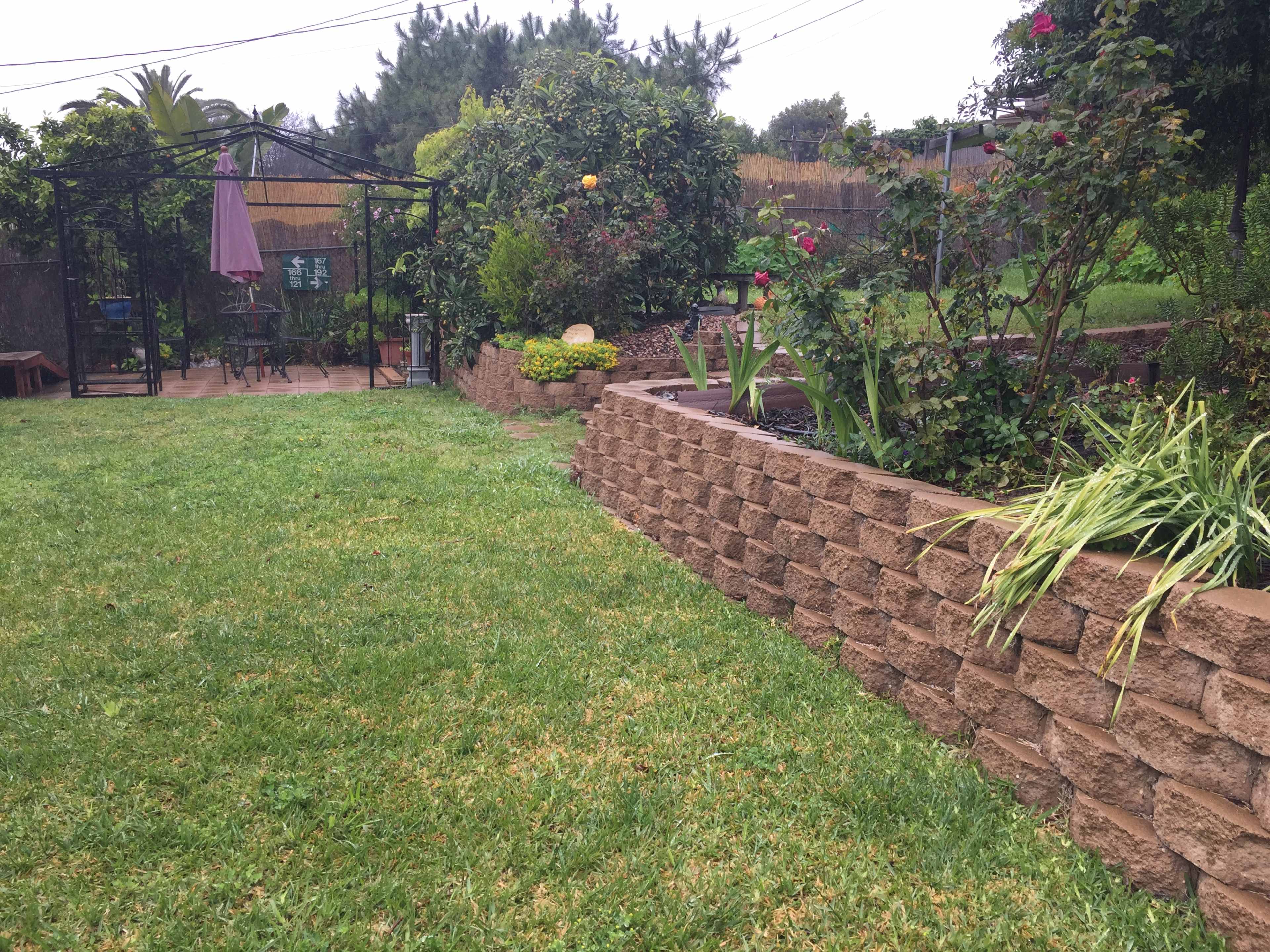 A landscaped backyard featuring a grassy area, a stone wall with plants, and a gazebo in the background.