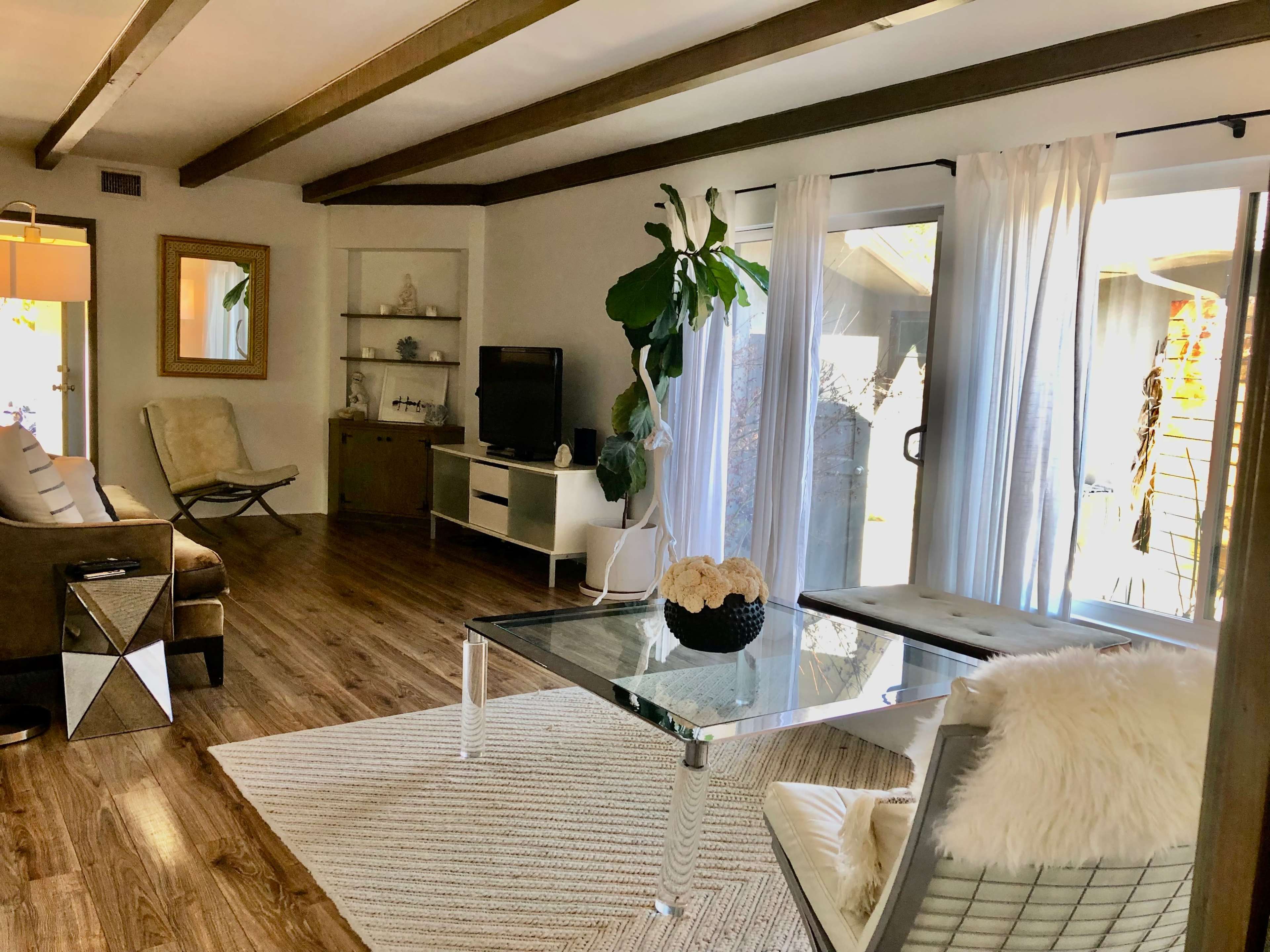 A spacious living room with wooden beams, a glass coffee table, a plush seating area, and large windows letting in natural light.