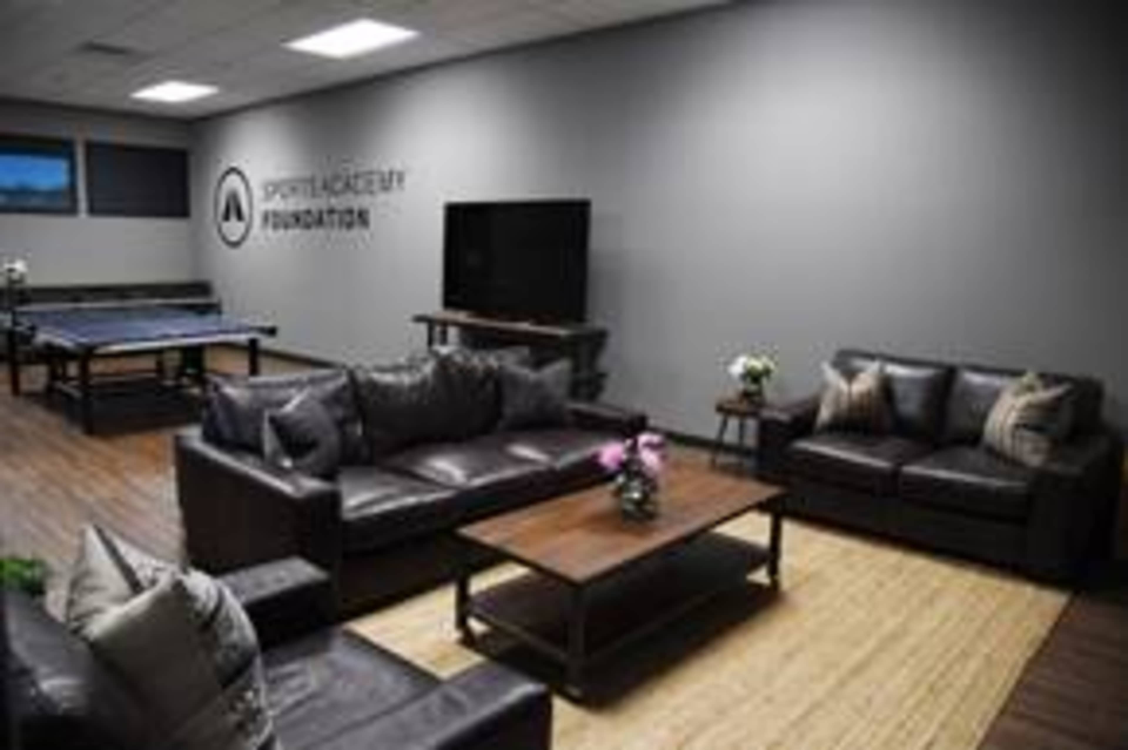 A modern lounge area with leather sofas, a coffee table, and a television, featuring the logo of the Sports Academy Foundation on the wall.