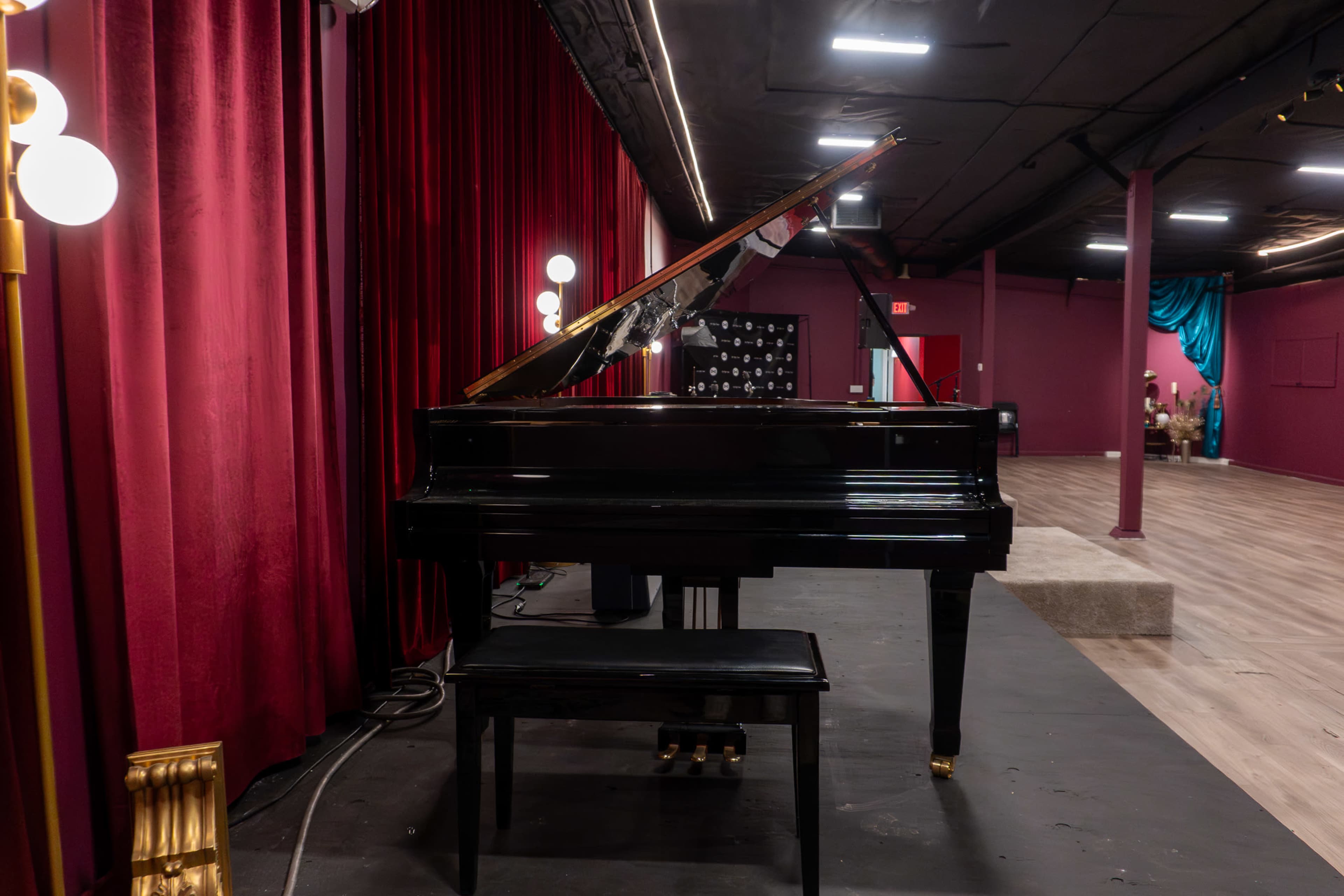A black grand piano is positioned on a stage in front of red curtains, with a wooden bench nearby and a backdrop of dim lighting.