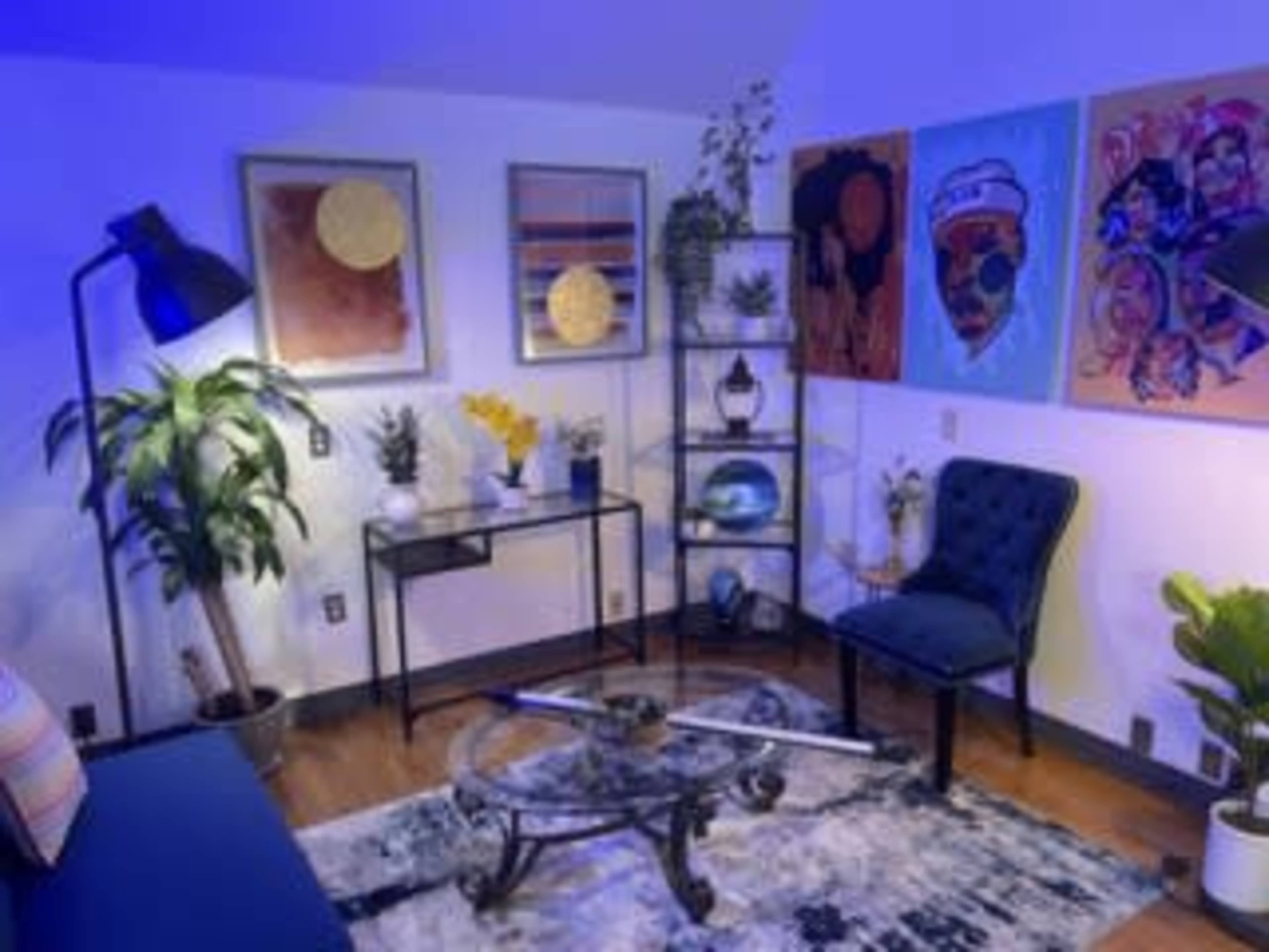 A cozy room with a seating area featuring a blue chair and a glass coffee table, adorned with plants and colorful artwork on the walls.