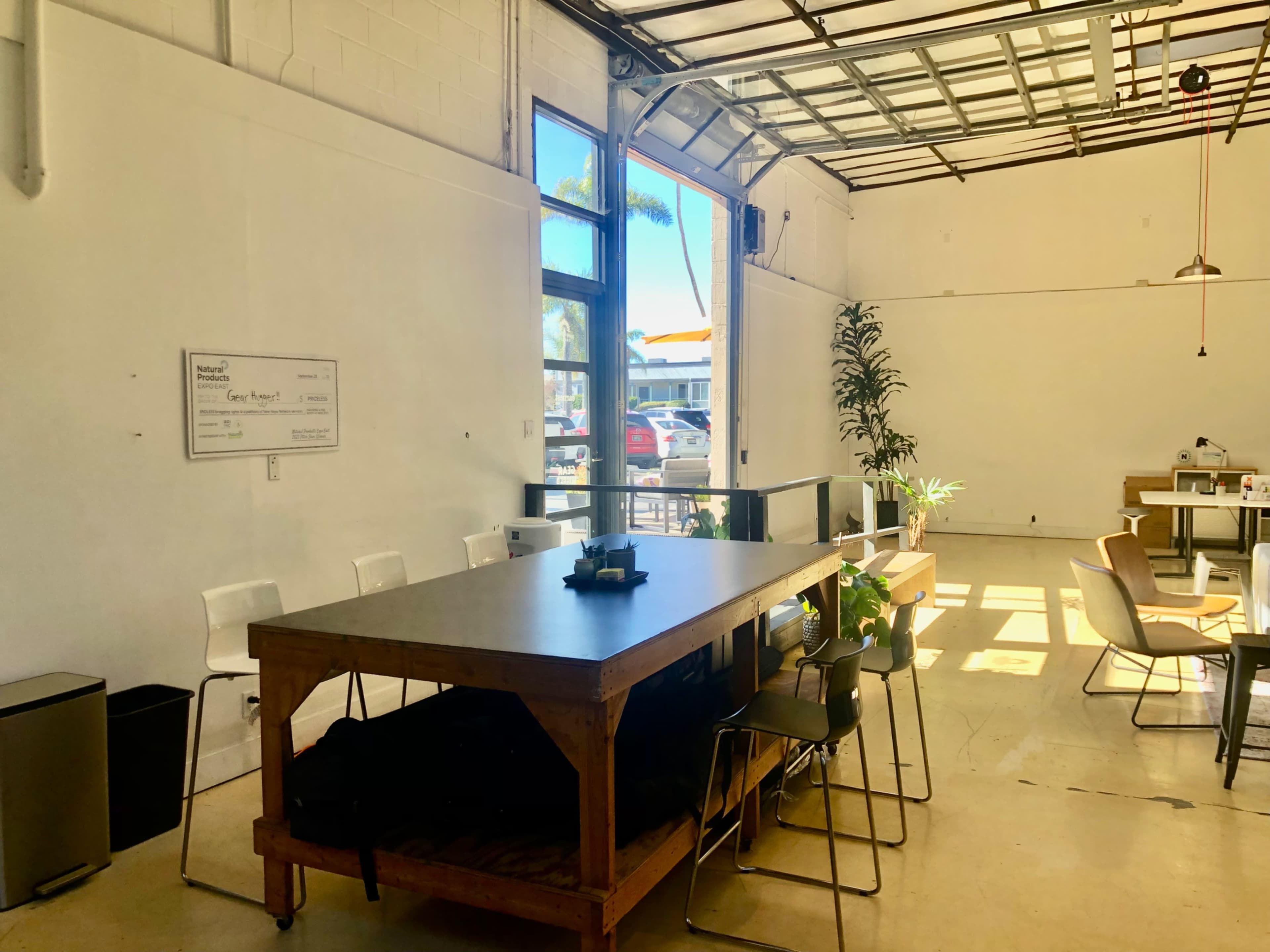 A spacious room features a large wooden table surrounded by chairs, with a waste bin visible in one corner and large windows allowing daylight to fill the space.