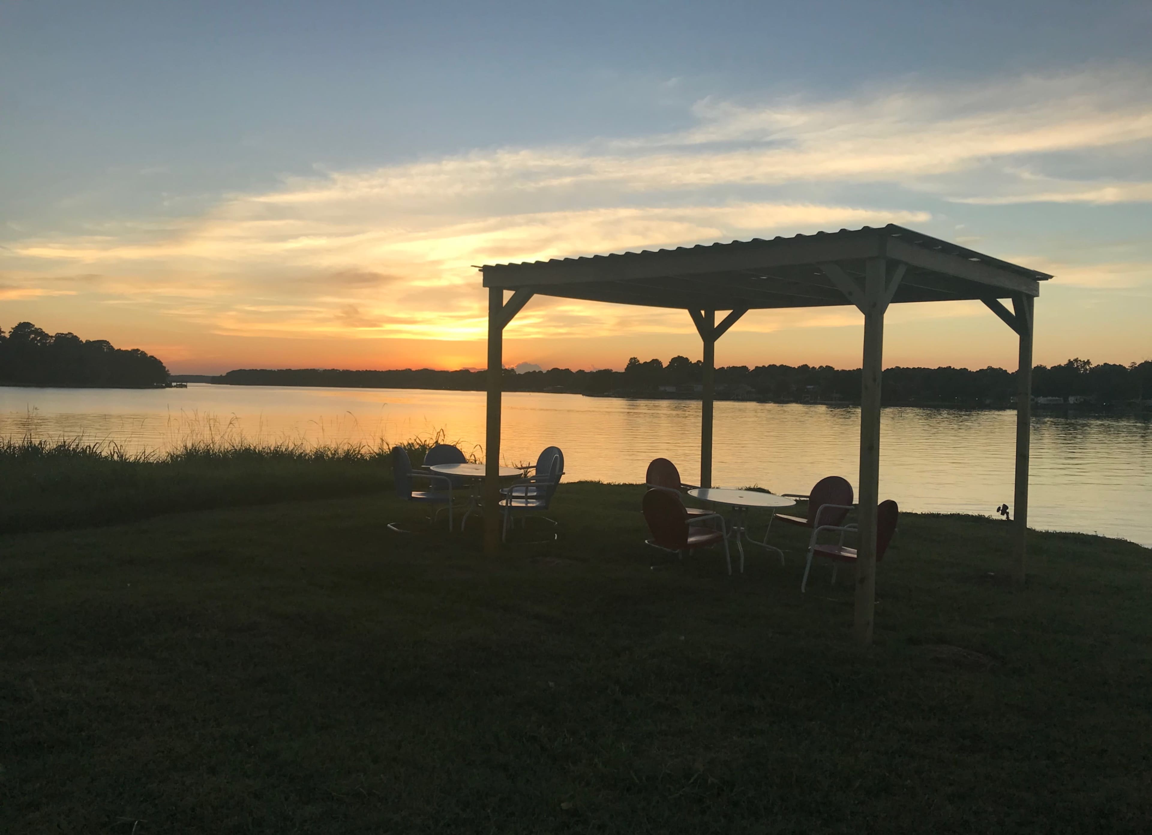 A gazebo with chairs and tables overlooks a calm lake at sunset.