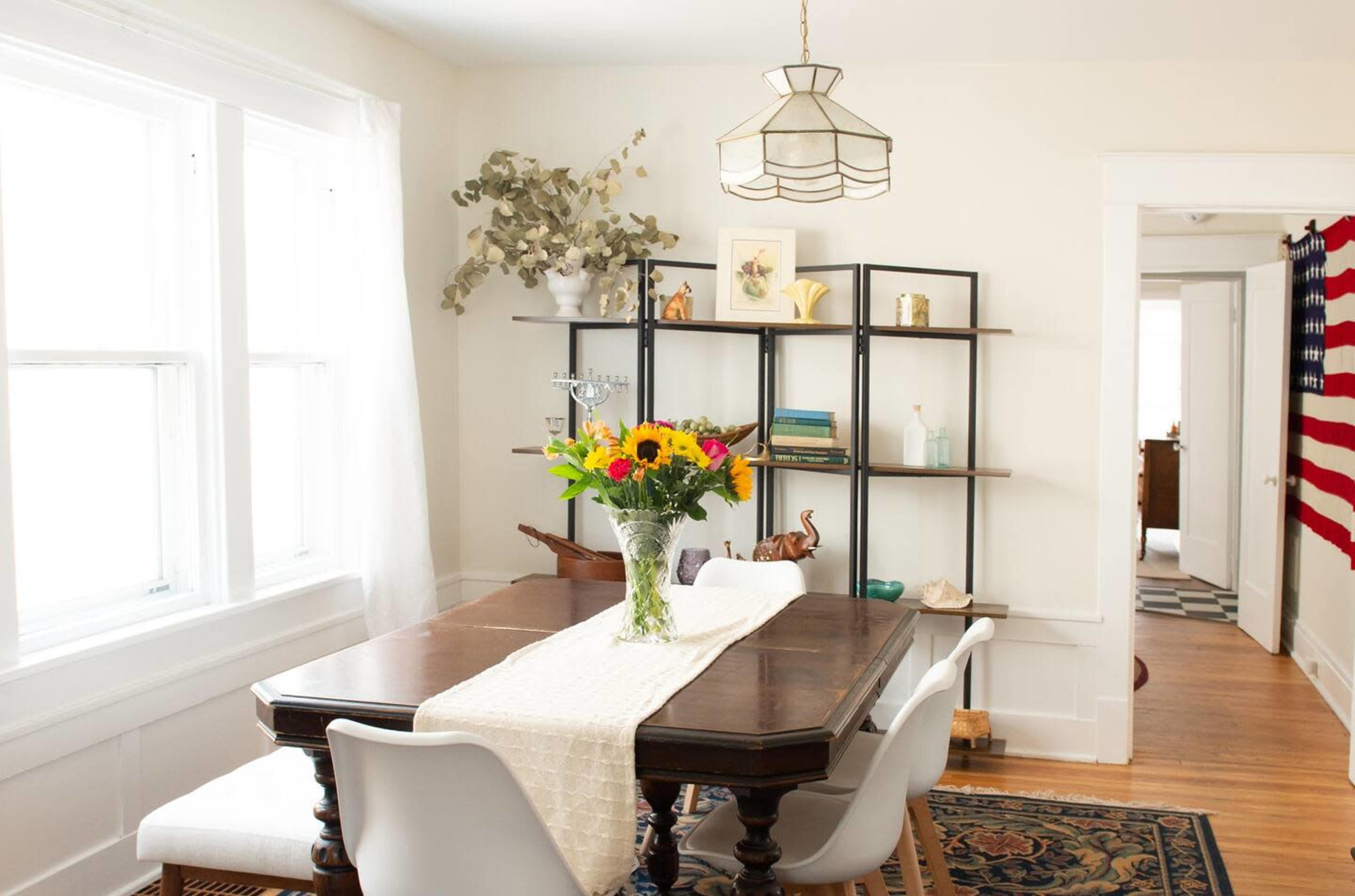 A dining area features a wooden table with a floral centerpiece, surrounded by white chairs, and a shelf displaying various decorative items, with a flag hanging in the background.