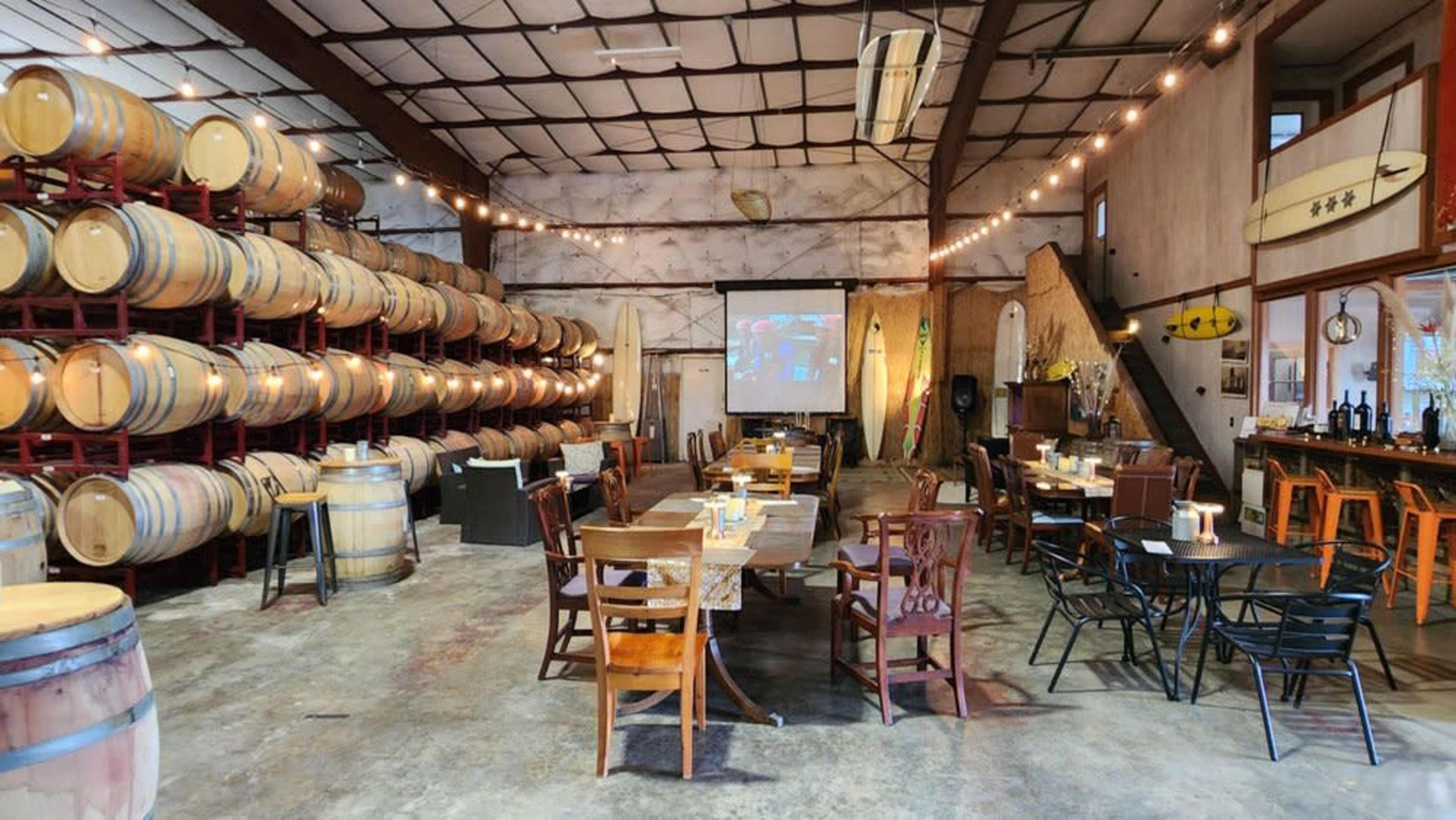 The image shows a rustic winery interior with stacked wine barrels, tables set for dining, and a projector screen in the background.