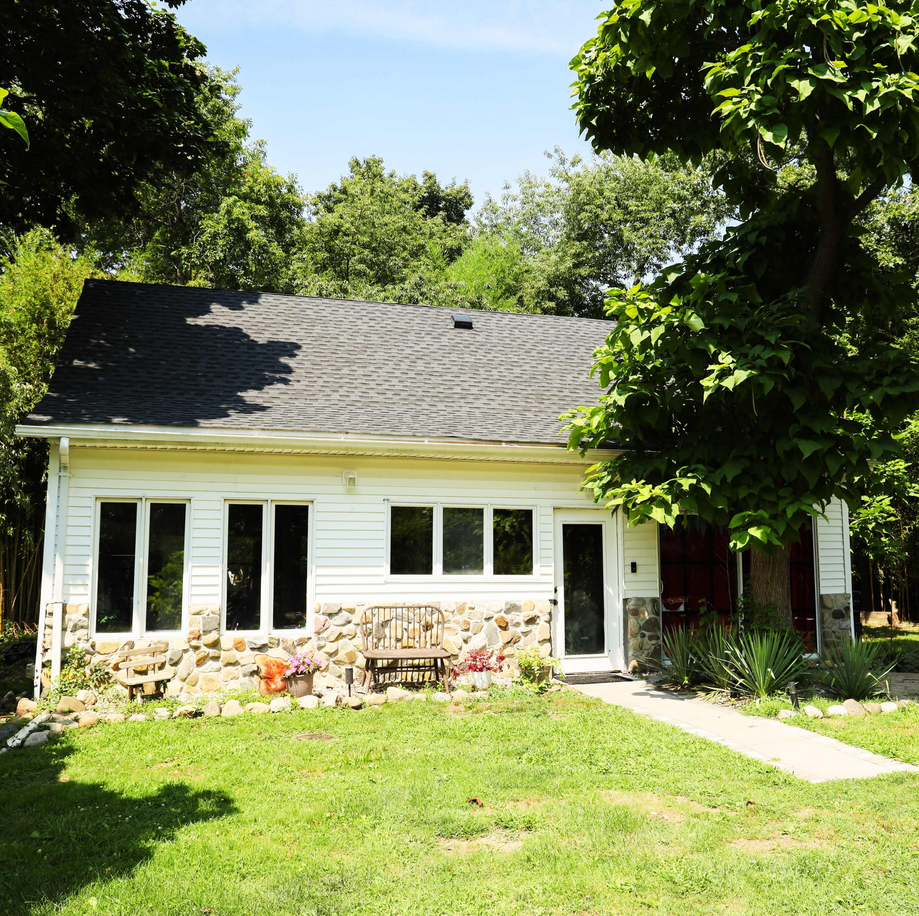 A single-story house with a stone facade and a wooden bench sits in a grassy area surrounded by trees.