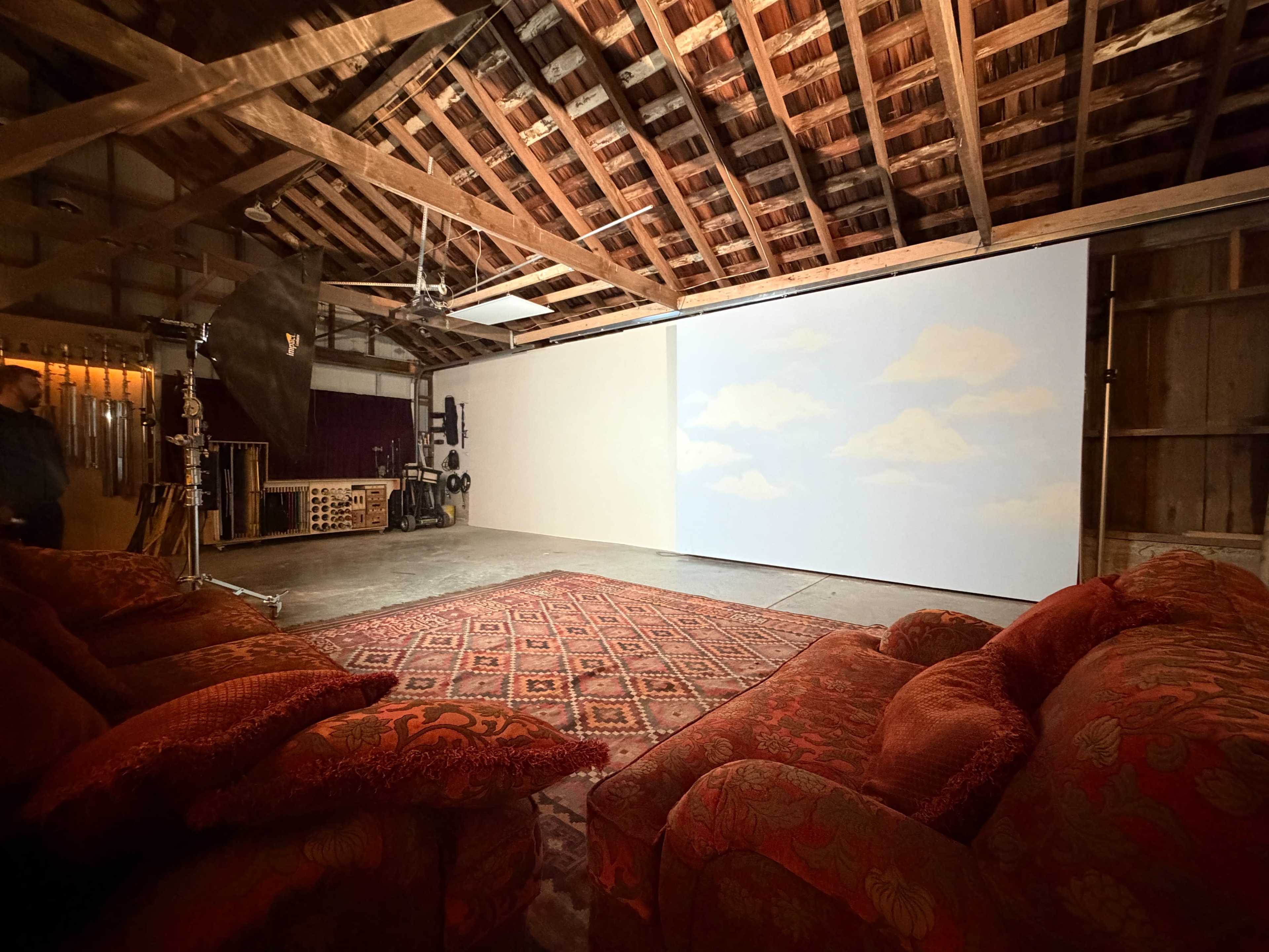 The image shows an interior space with a wooden beam ceiling, featuring a large projection screen displaying a sky scene, surrounded by a patterned rug and red upholstered sofas.
