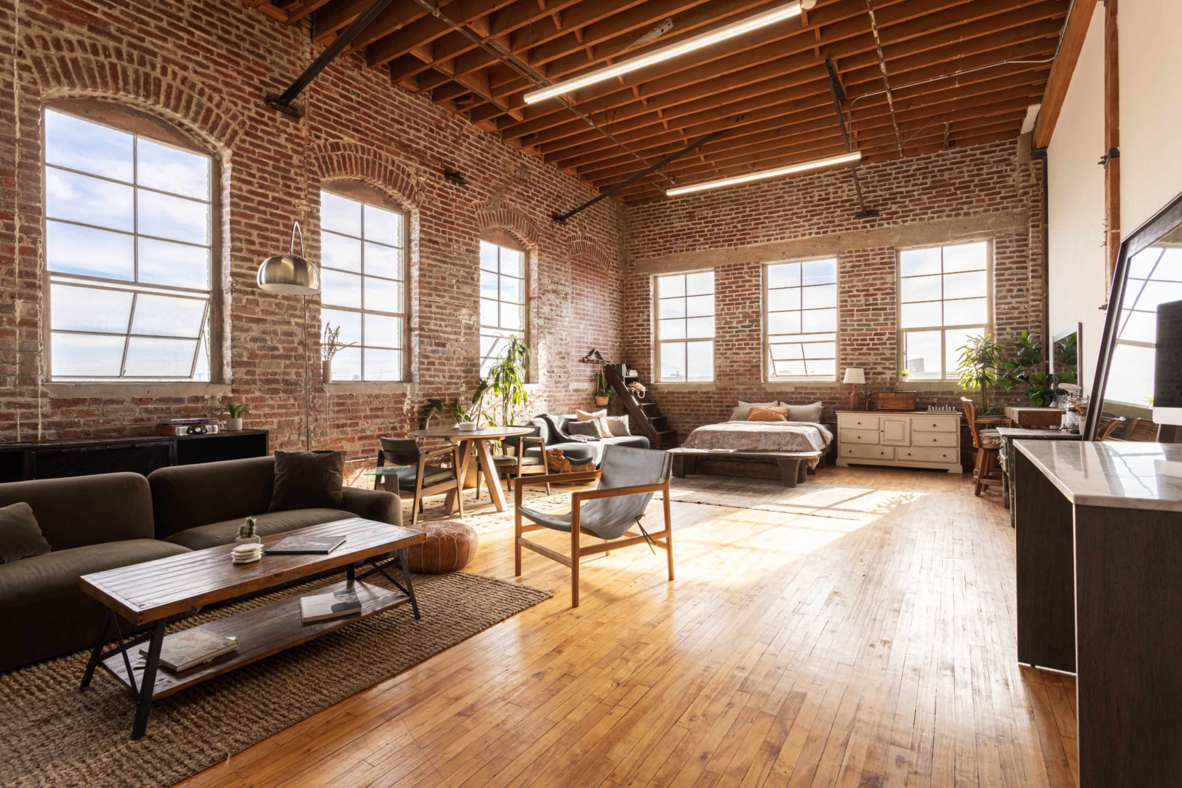 An industrial-style loft with exposed brick walls, large windows, wooden floors, and a blend of living and sleeping areas furnished with a sofa, a coffee table, and a bed.