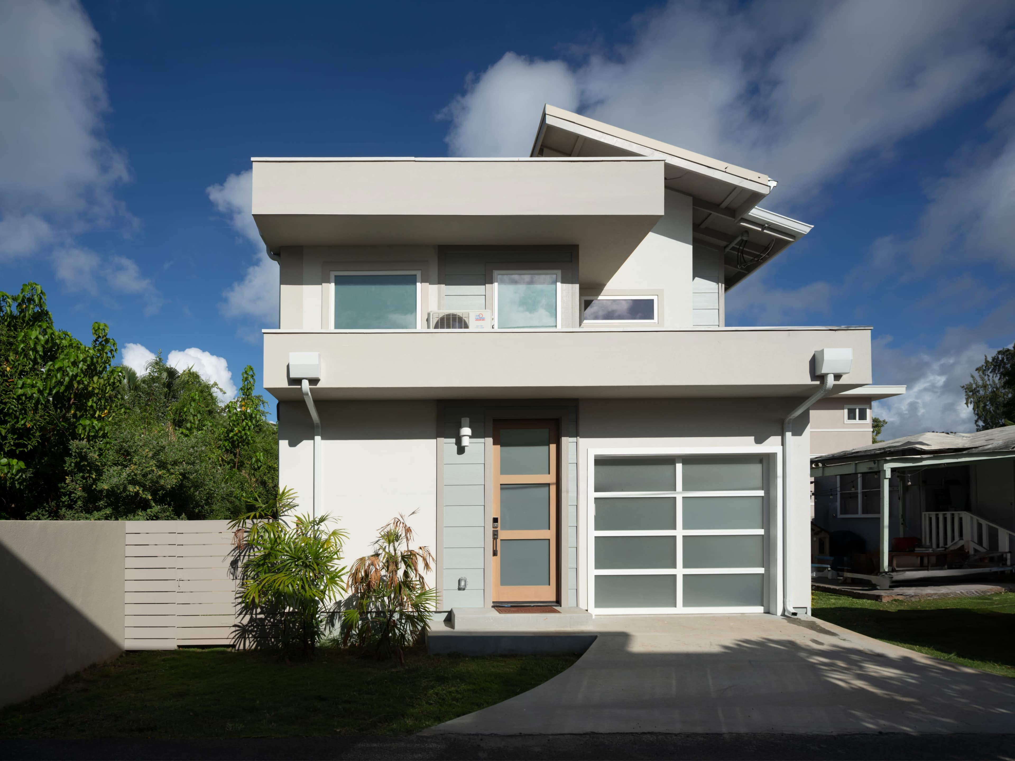 The image shows a modern two-story house with a flat roof and a garage door facing a driveway, surrounded by green trees and a clear blue sky.