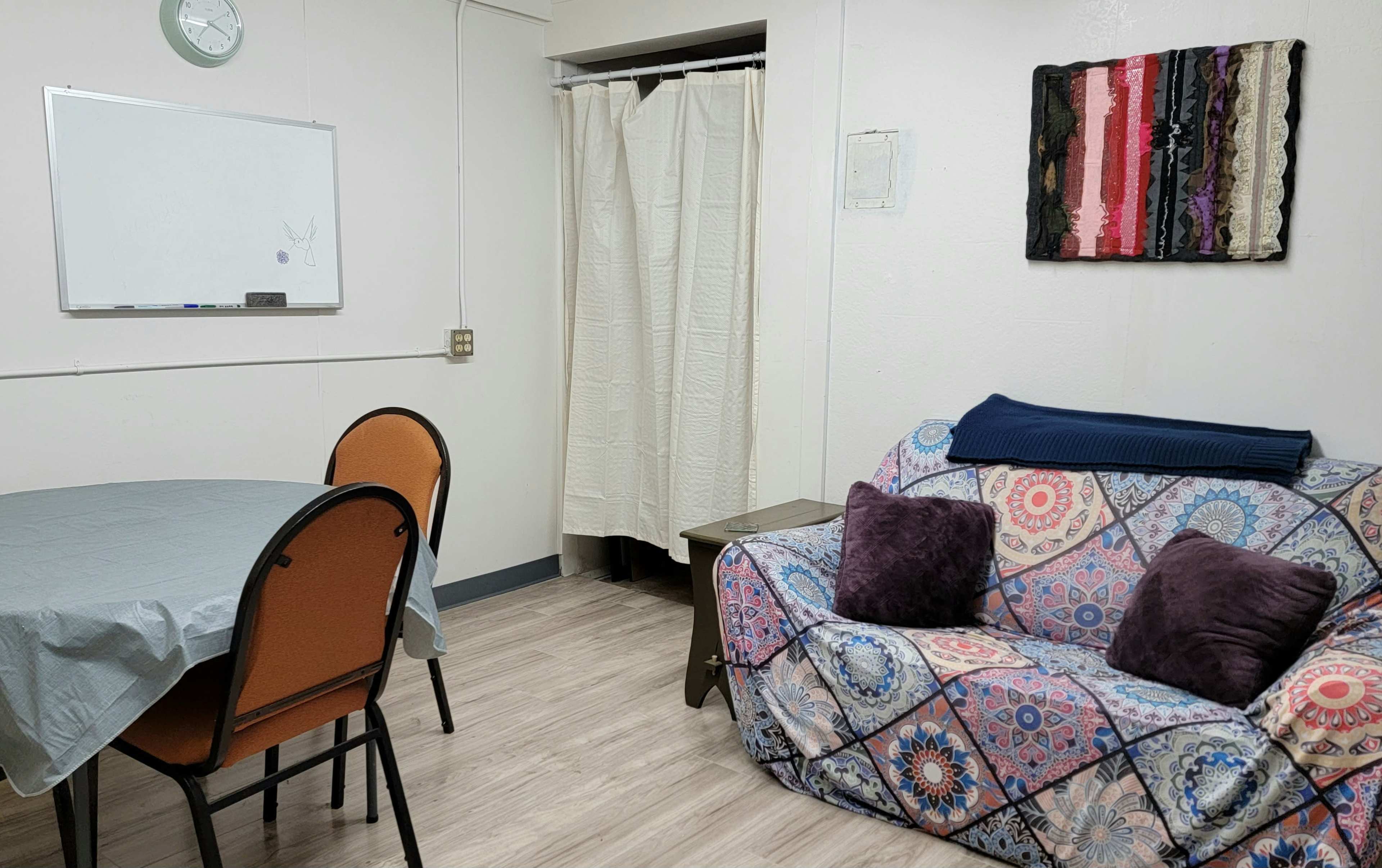 The image shows a small room with a table and four chairs, a couch covered in patterned fabric, and a whiteboard mounted on the wall.