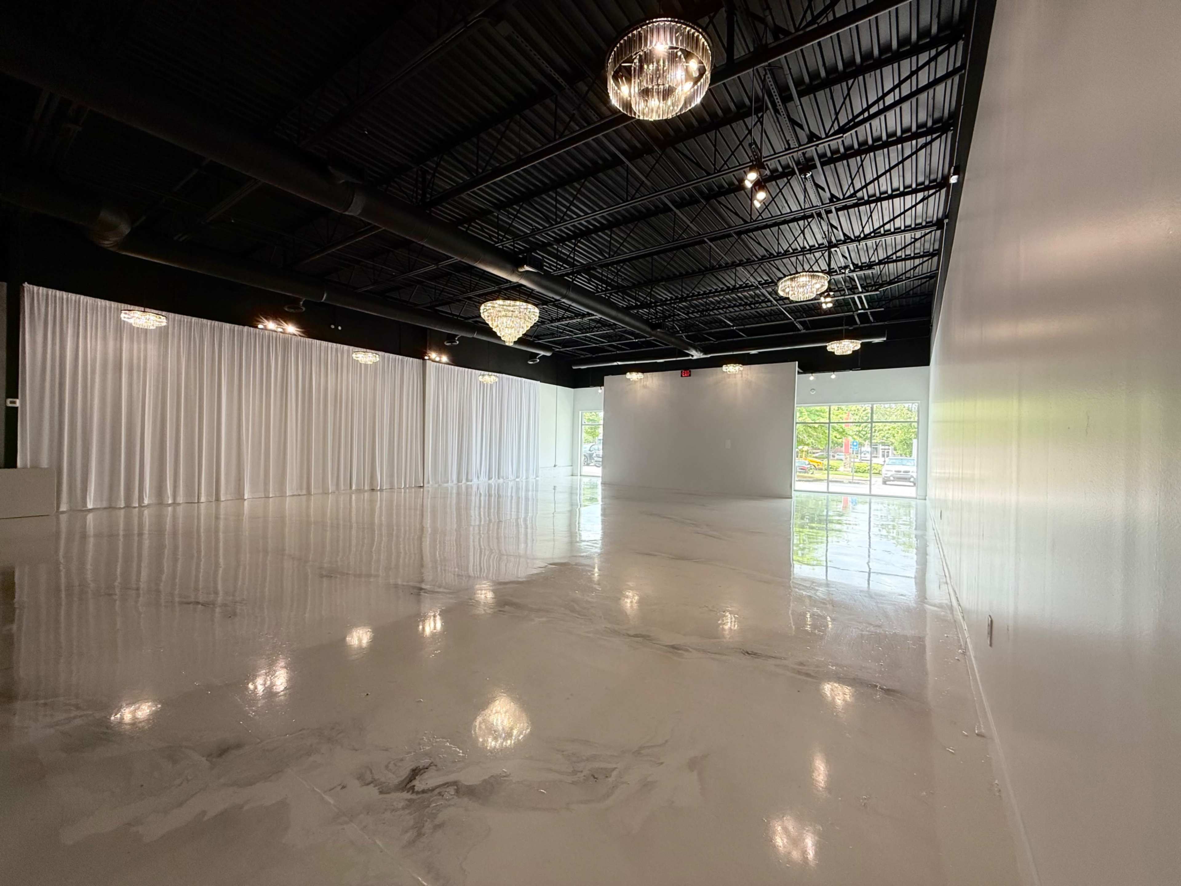 The image shows a spacious, empty room with polished concrete floors, white walls, and large windows letting in natural light, adorned with hanging chandeliers and sheer curtains.