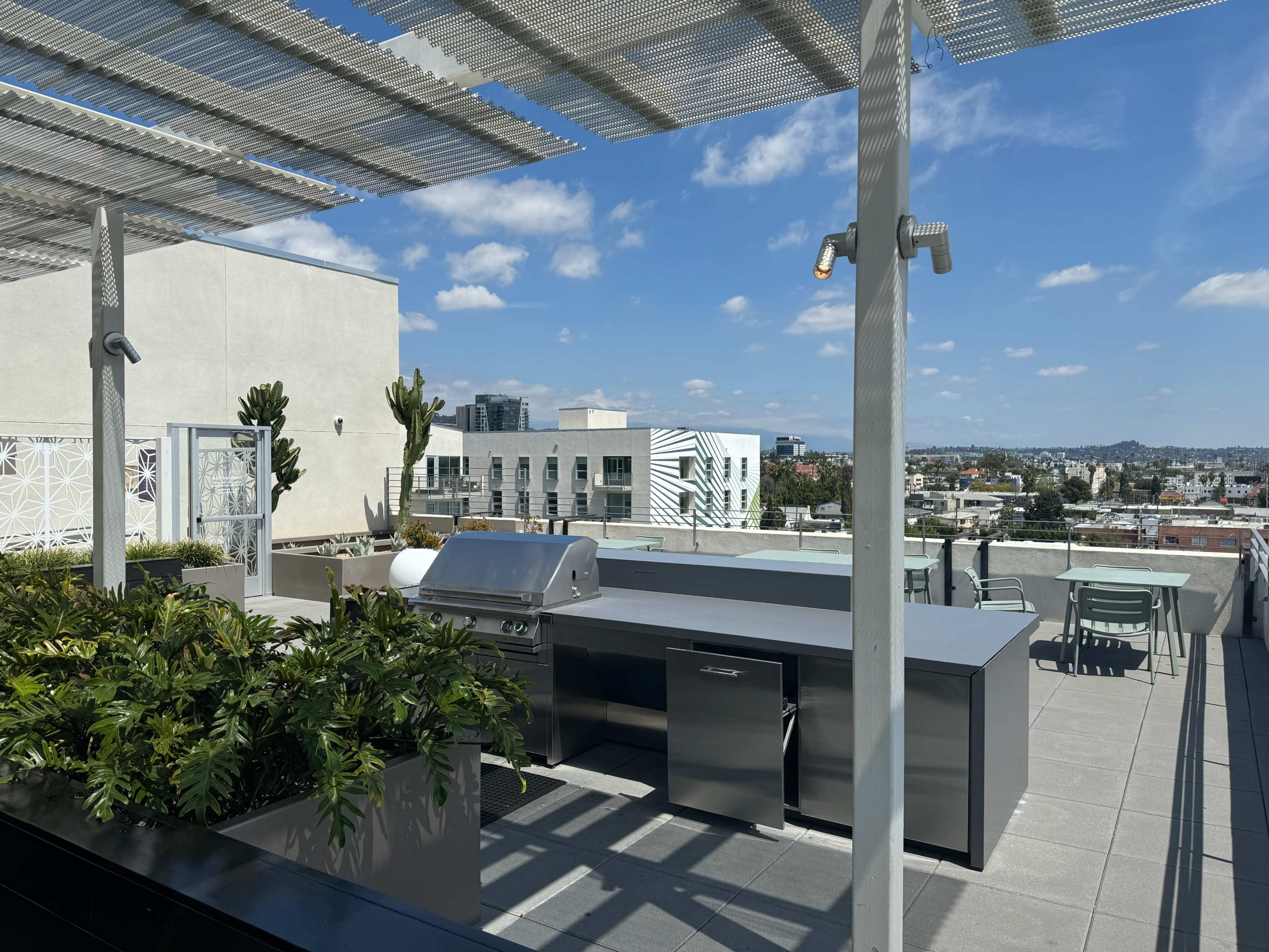 A stylish rooftop kitchen equipped with a grill and surrounded by greenery, set against a backdrop of a city skyline under a partly cloudy sky.
