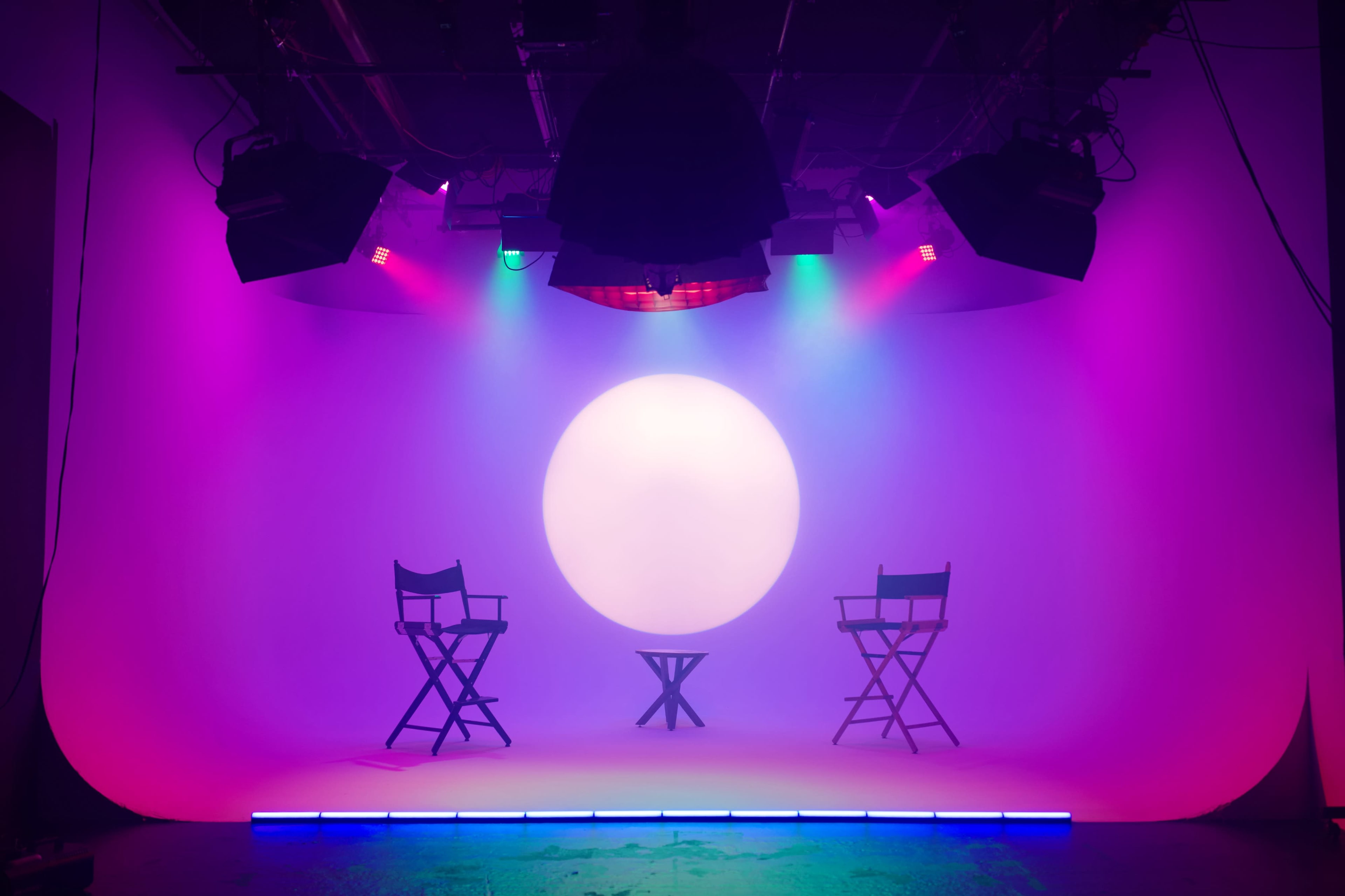 A photography studio features two director's chairs placed facing a small round table under vibrant colored lights with a large illuminated circle in the background.