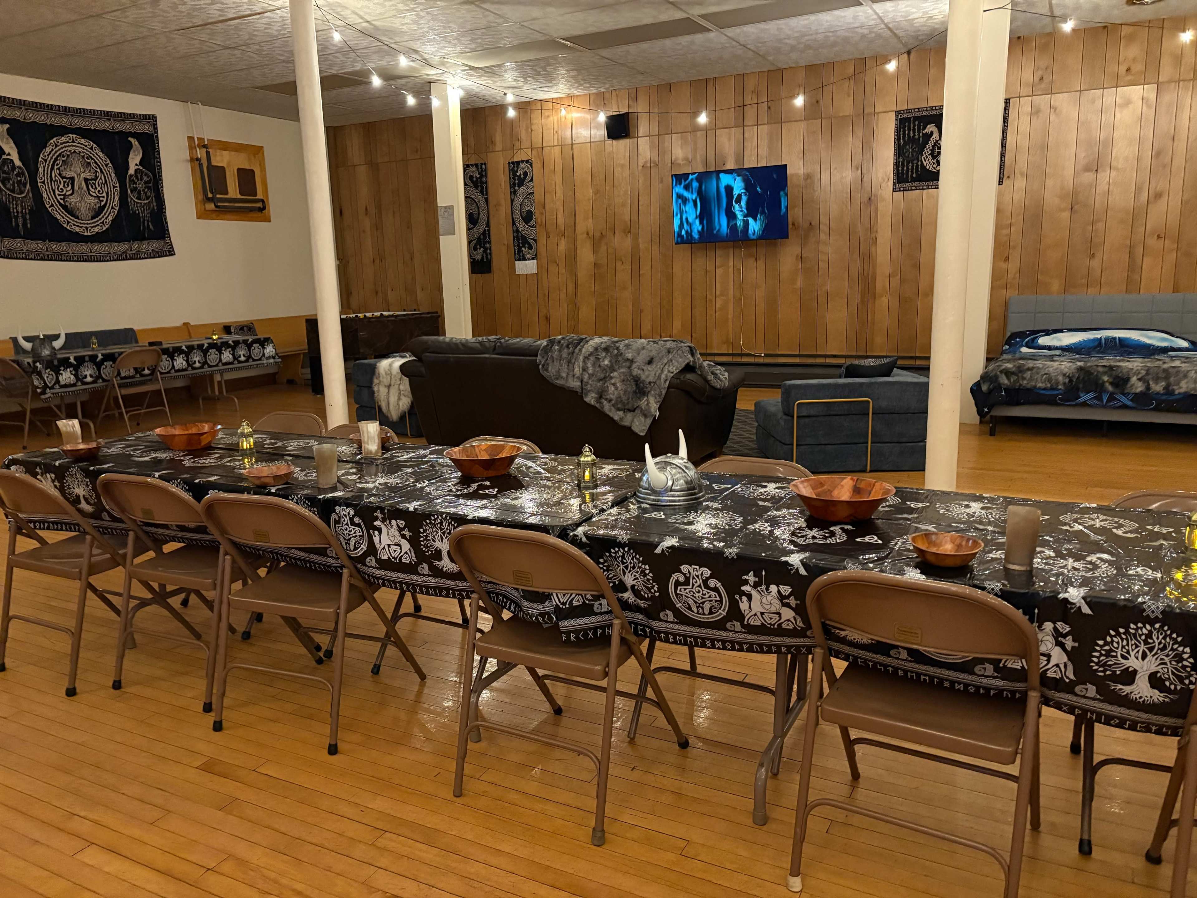 The image shows a spacious room with wooden paneling, featuring long tables set with dark tablecloths, chairs, and various decorative bowls, along with a sofa area and a television on the wall.
